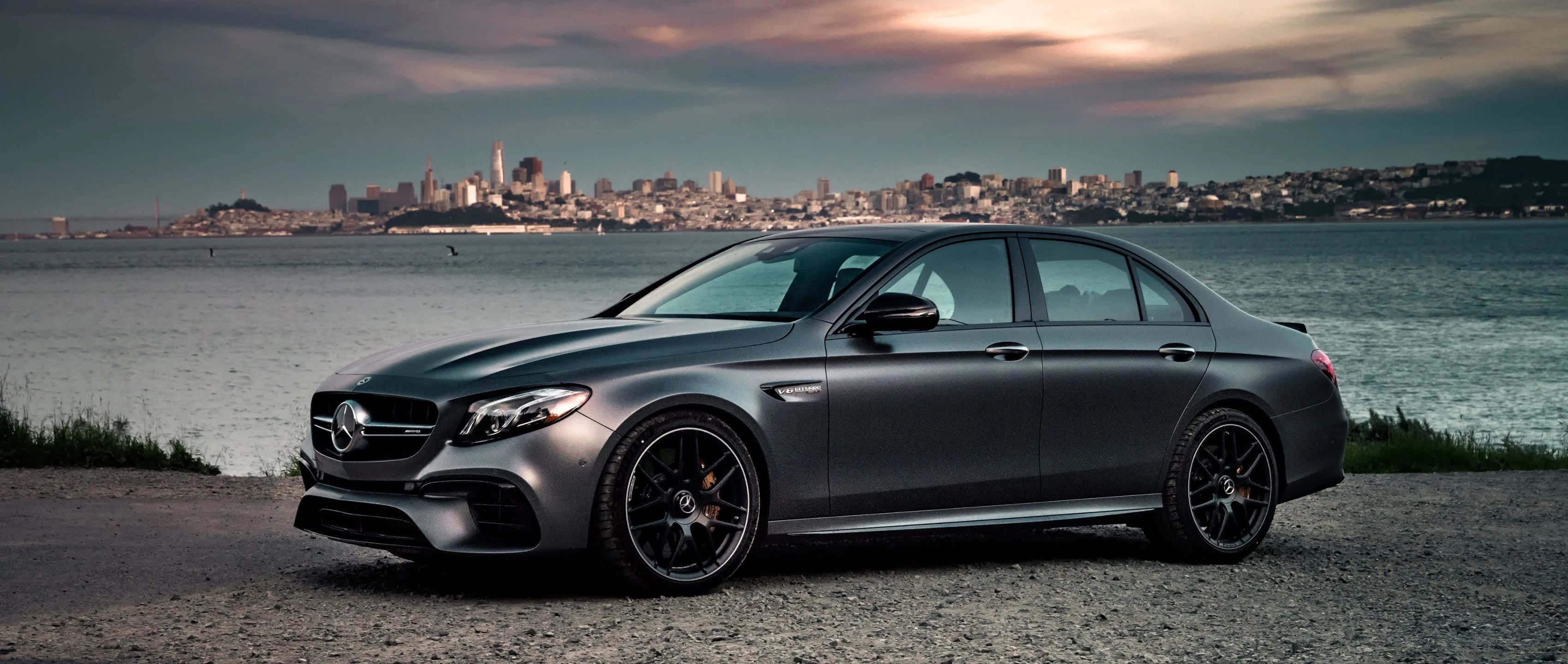 Coupe 桌布 壁紙 — 一輛啞光灰色的梅賽德斯-AMG E63轎車停在礫石岸邊，水對岸的城市天際線在陰沉的暮色天空下。