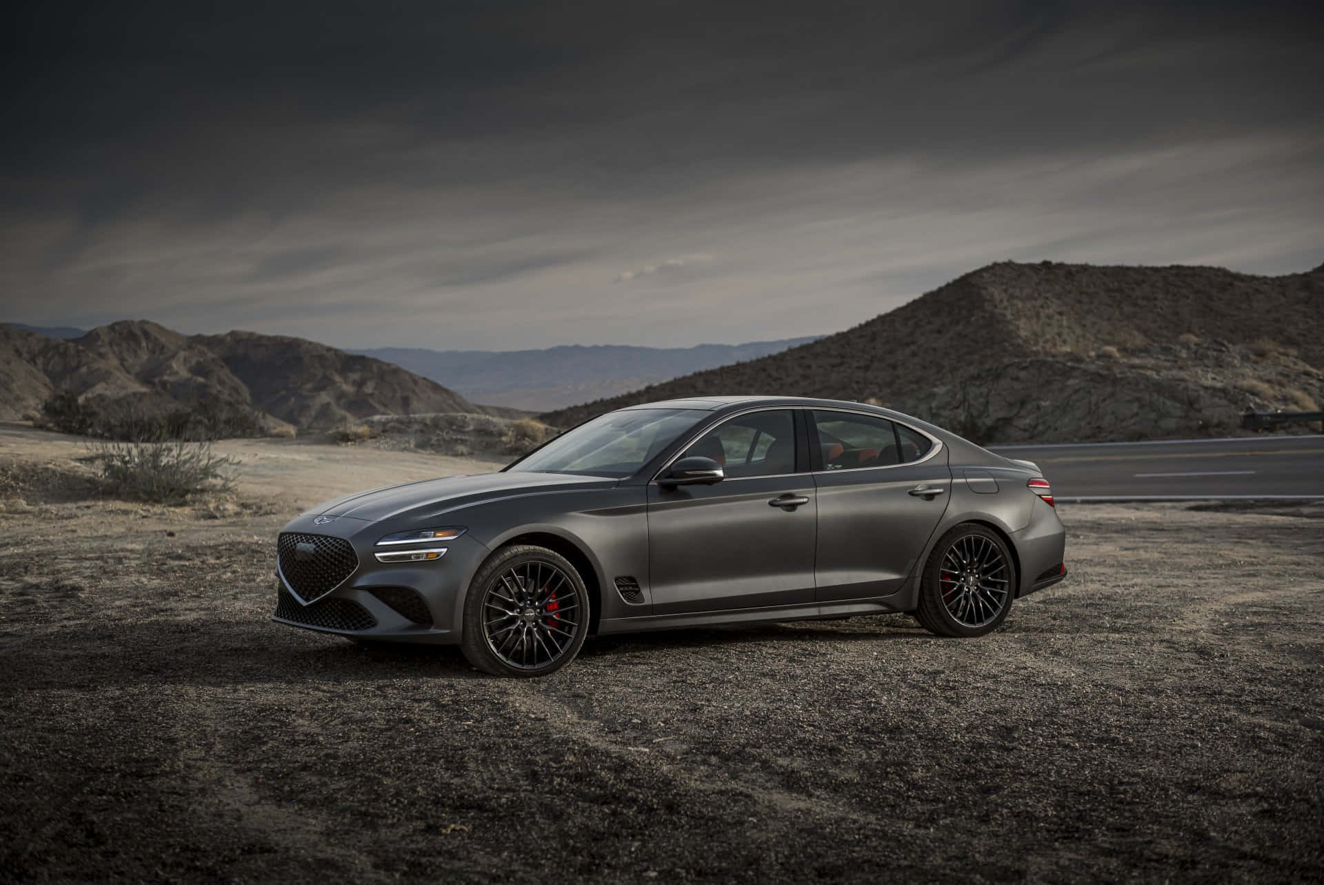 Genesis G70 桌布 壁紙 — 一輛時尚的啞光灰色運動轎車配黑色輪圈，停在碎石沙漠道路上，天空陰沉多雲，遠處有山丘。