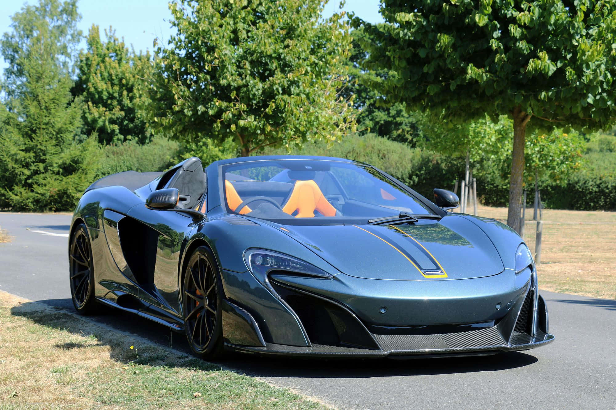 Mclaren 675lt Spider 桌布 壁紙 — 一輛時尚的啞光灰色McLaren跑車，配橙色座椅，停泊在陽光普照的道路旁，旁邊是茂密的綠樹。