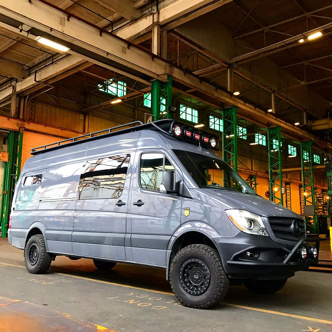 Mercedes Benz Sprinter 桌布 壁紙 — 一輛啞光灰色的 Mercedes-Benz Sprinter 客貨車，配備堅固輪胎和車頂架，停放在高天花板的工業倉庫內。