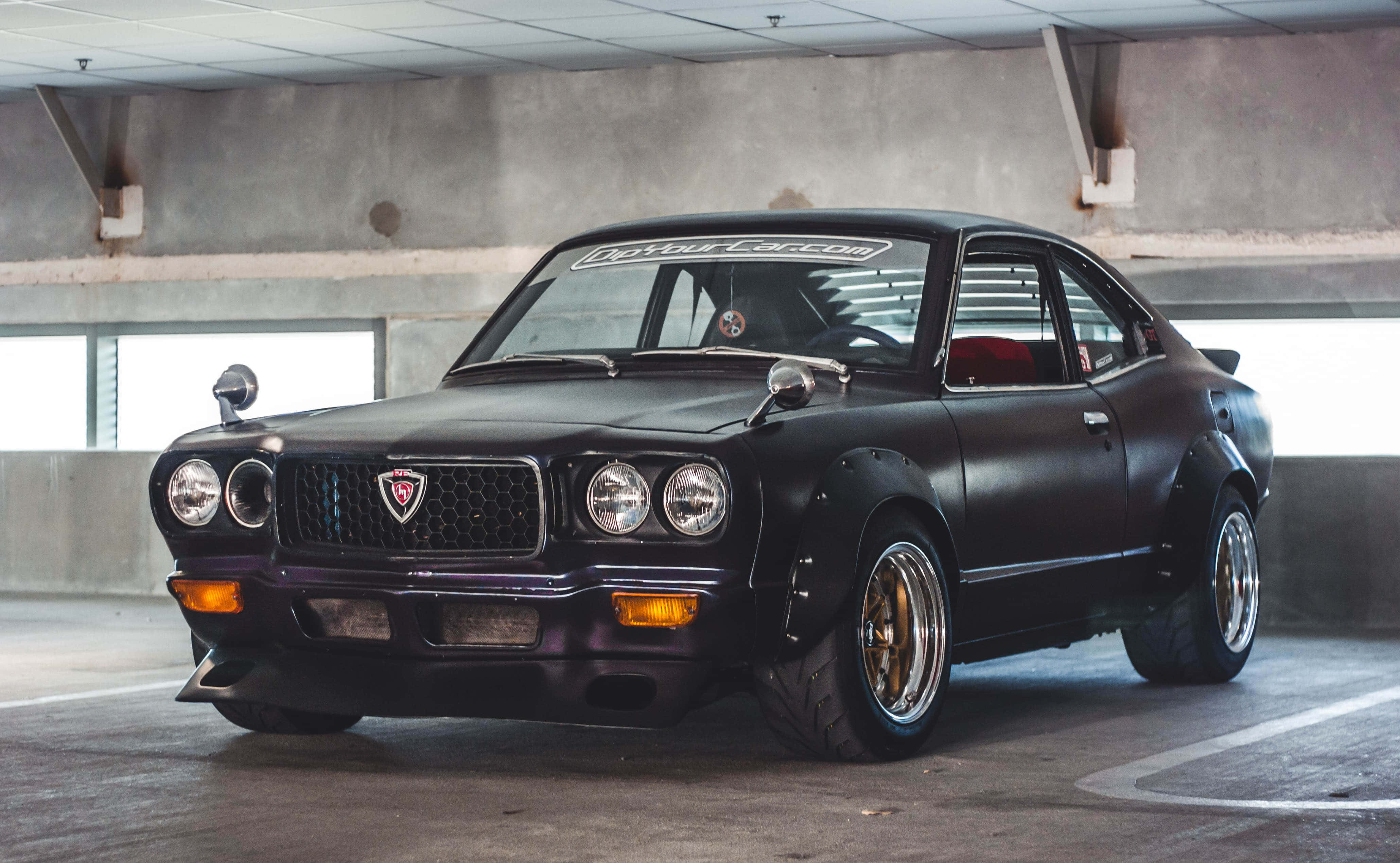 Mazda Rx-3 桌布 壁紙 — 一輛啞光黑色經典日本雙門跑車配金色輪圈停泊在混凝土停車場。