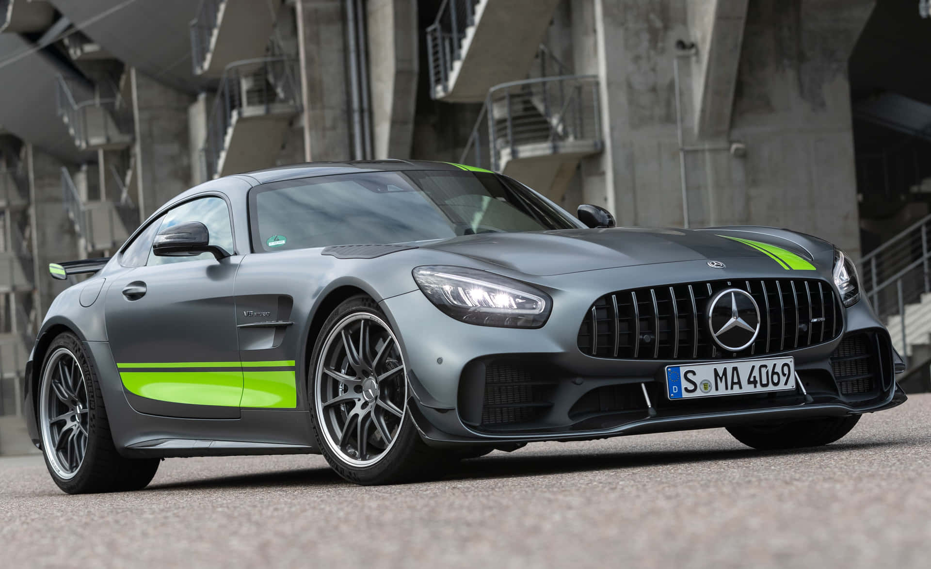  壁紙 — 一輛啞光灰色的梅賽德斯-AMG GT R 跑車，帶有螢光綠色條紋，停在混凝土體育場建築前。