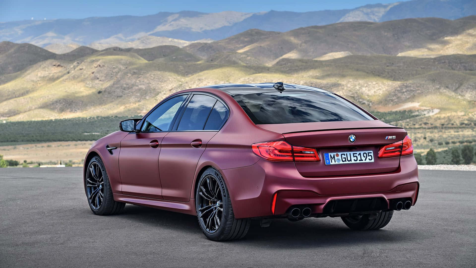 Bmw M5 桌布 壁紙 — 一輛啞光紅色BMW M5房車停在瀝青路上，背景是起伏的丘陵和遠處的山脈，天空清澈湛藍。