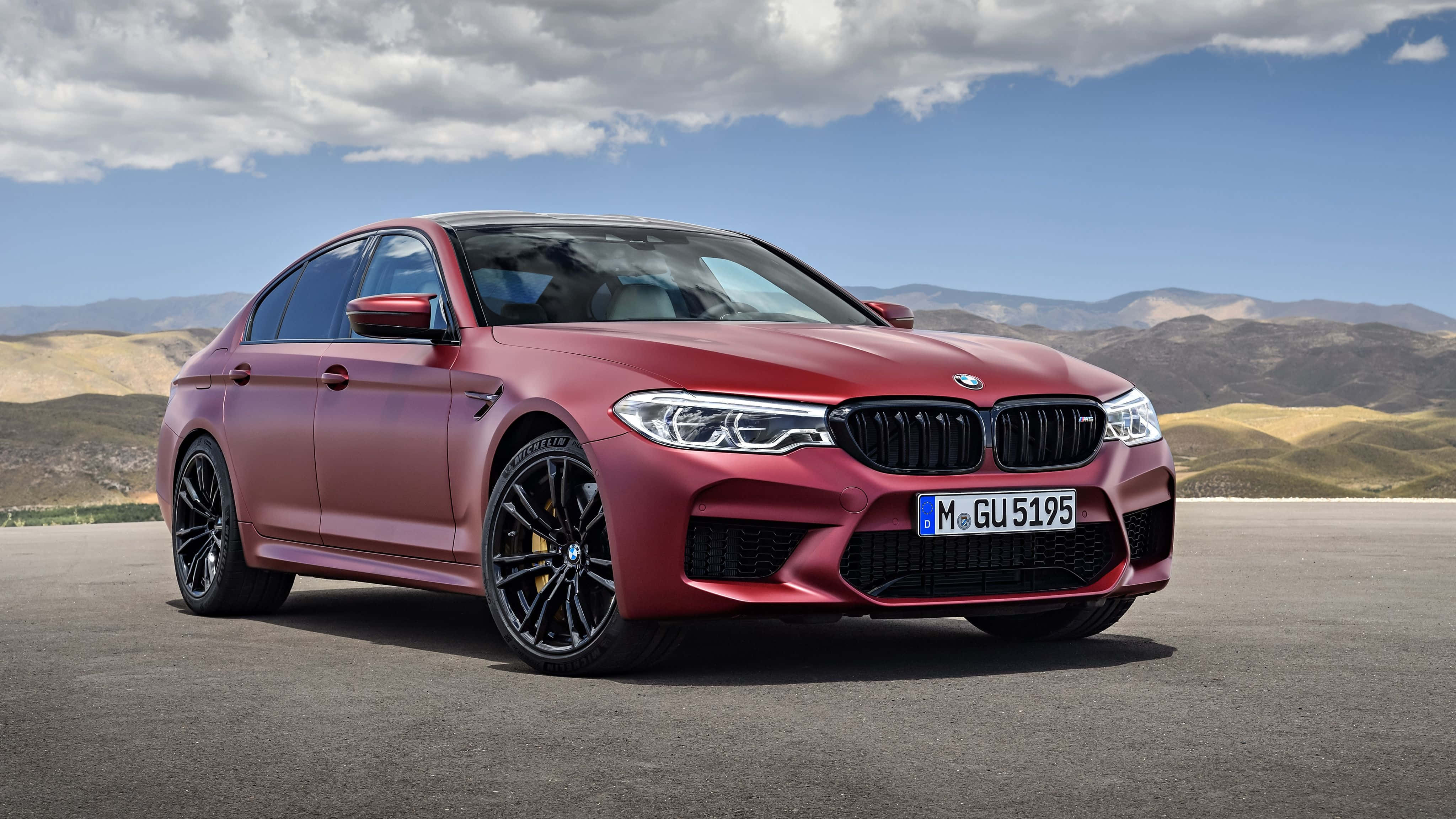 Bmw M5 桌布 壁紙 — 一架啞光紅色BMW M5房車停在瀝青路上，背景有山脈和陰天。