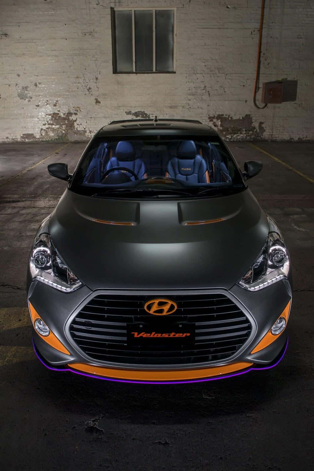 Hyundai Veloster 桌布 壁紙 — 一輛啞光黑色現代Veloster帶有橙色和紫色點綴，在昏暗的工業車庫中正面朝前。