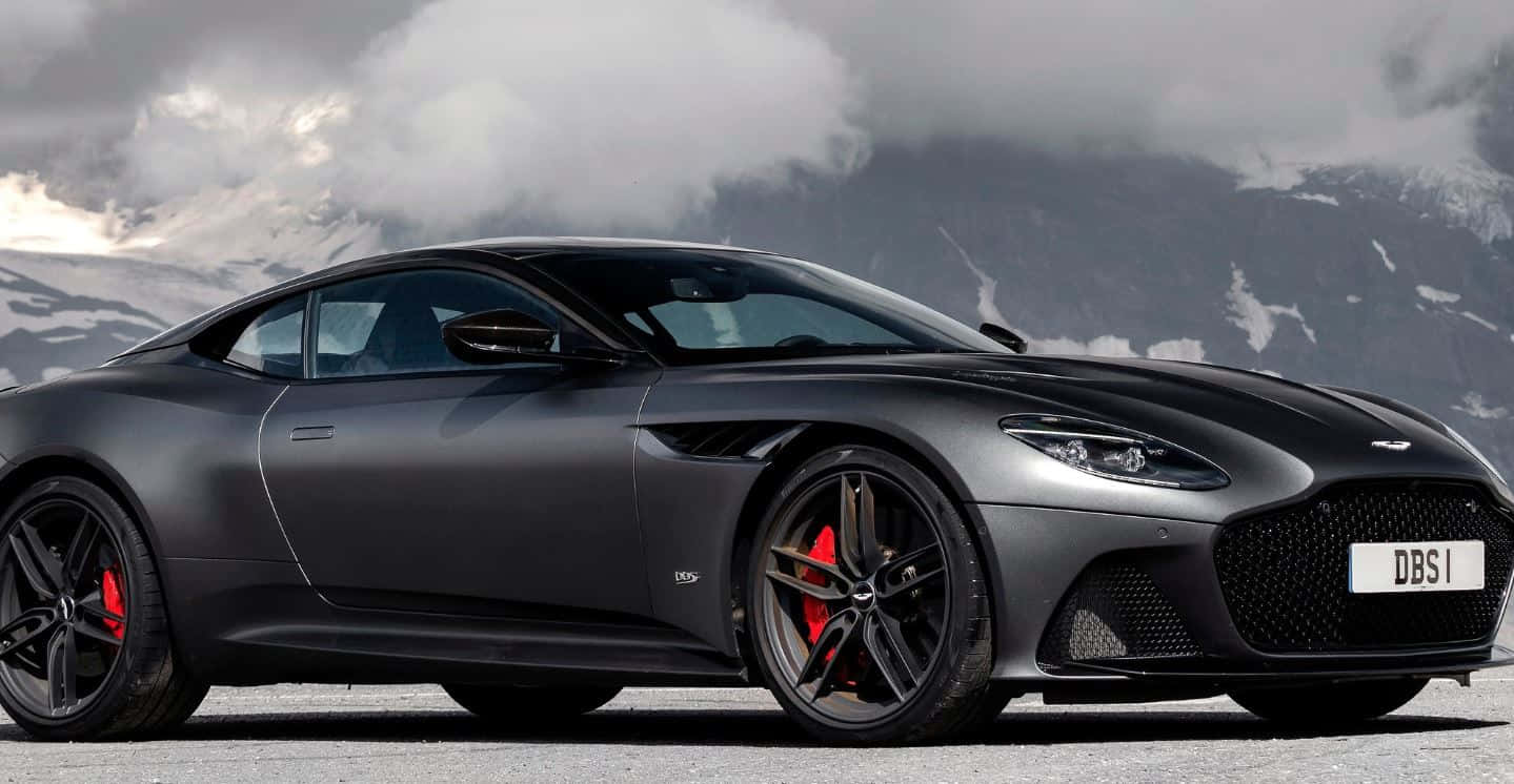 Aston Martin Dbs Superleggera 桌布 壁紙 — 一架型格啞黑色Aston Martin DBS1超級跑車，配紅色制動卡鉗，停泊在積雪的山路上，天空有戲劇性的雲層。