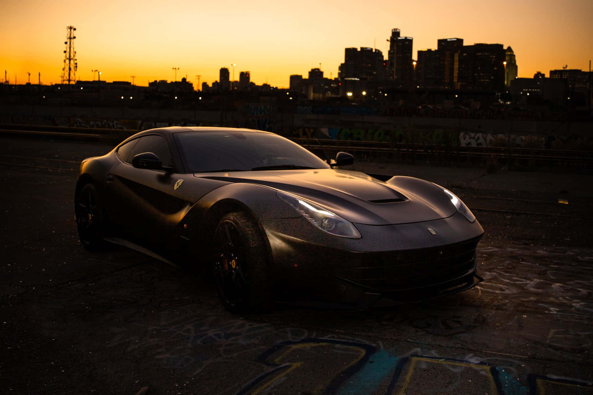 Ferrari F12 Berlinetta 桌布 壁紙 — 一架啞黑色法拉利F12 Berlinetta停泊在日落時分被塗鴉覆蓋的城市空地，背景是城市天際線。