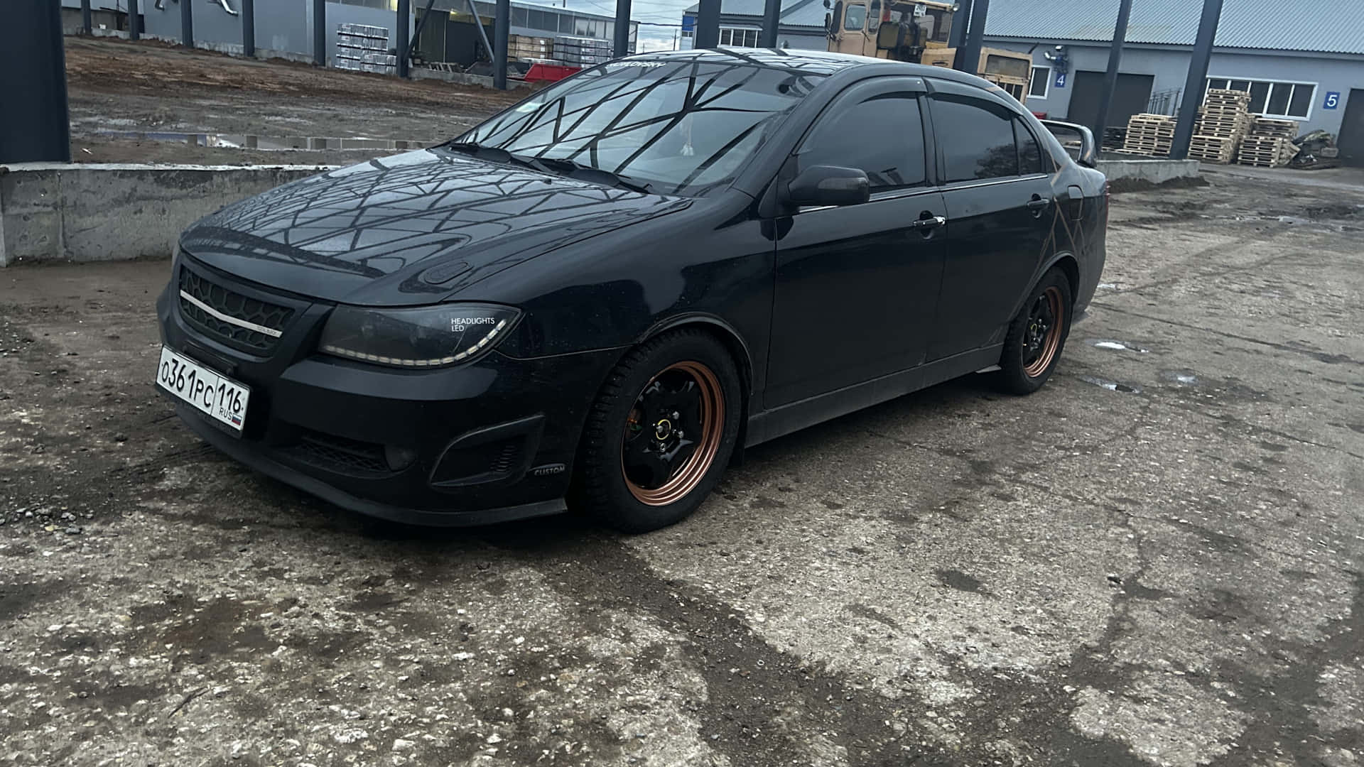  壁紙 — 一輛啞黑色改裝轎車配銅色輪圈，停在工業建築附近風化的混凝土場地上。