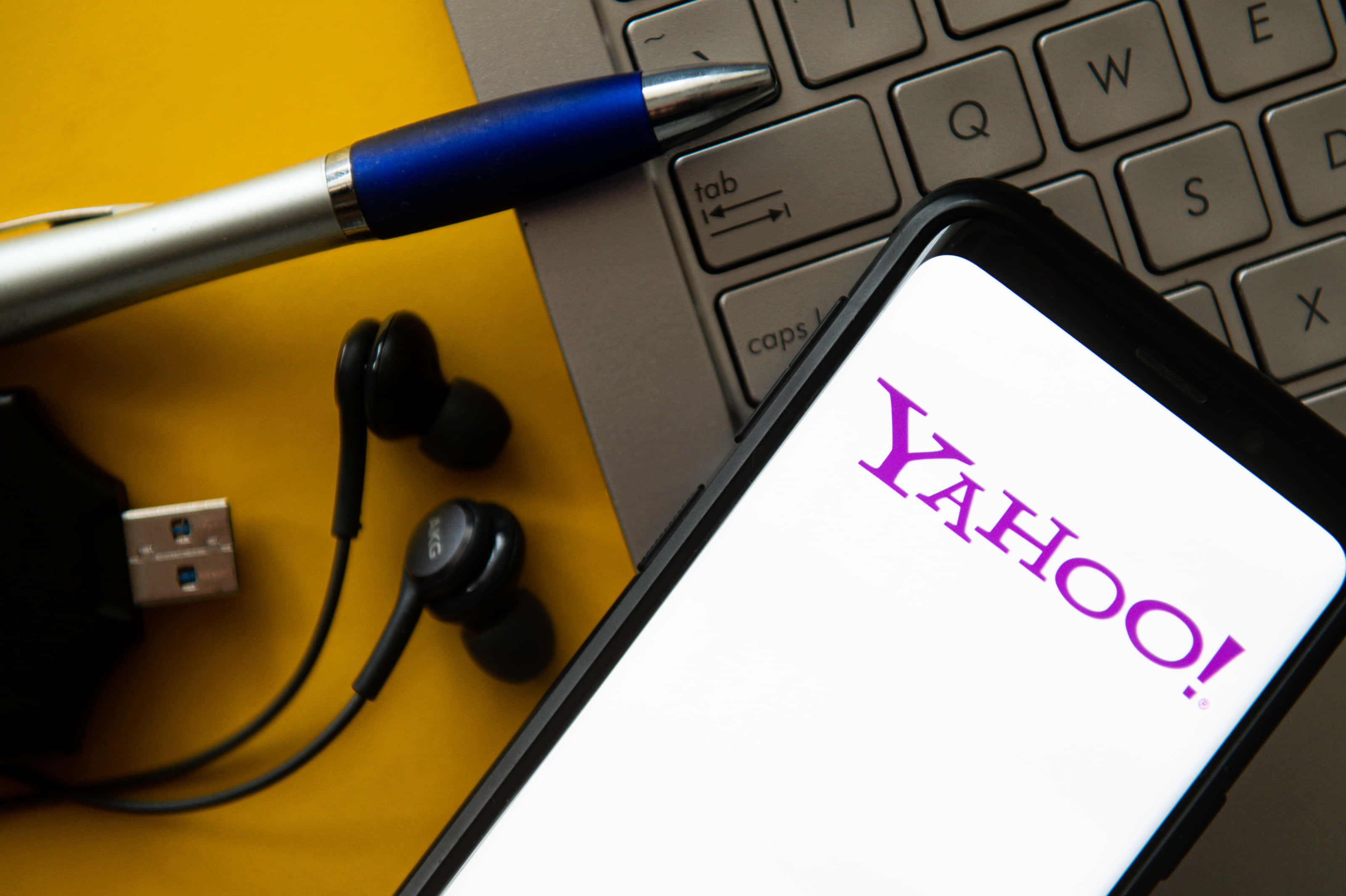 Yahoo! 多元與創新的產品線 壁紙 — 一部顯示Yahoo標誌的智能手機放在一個鮮黃色表面上，旁邊有手提電腦鍵盤、藍色筆、耳塞和USB手指。