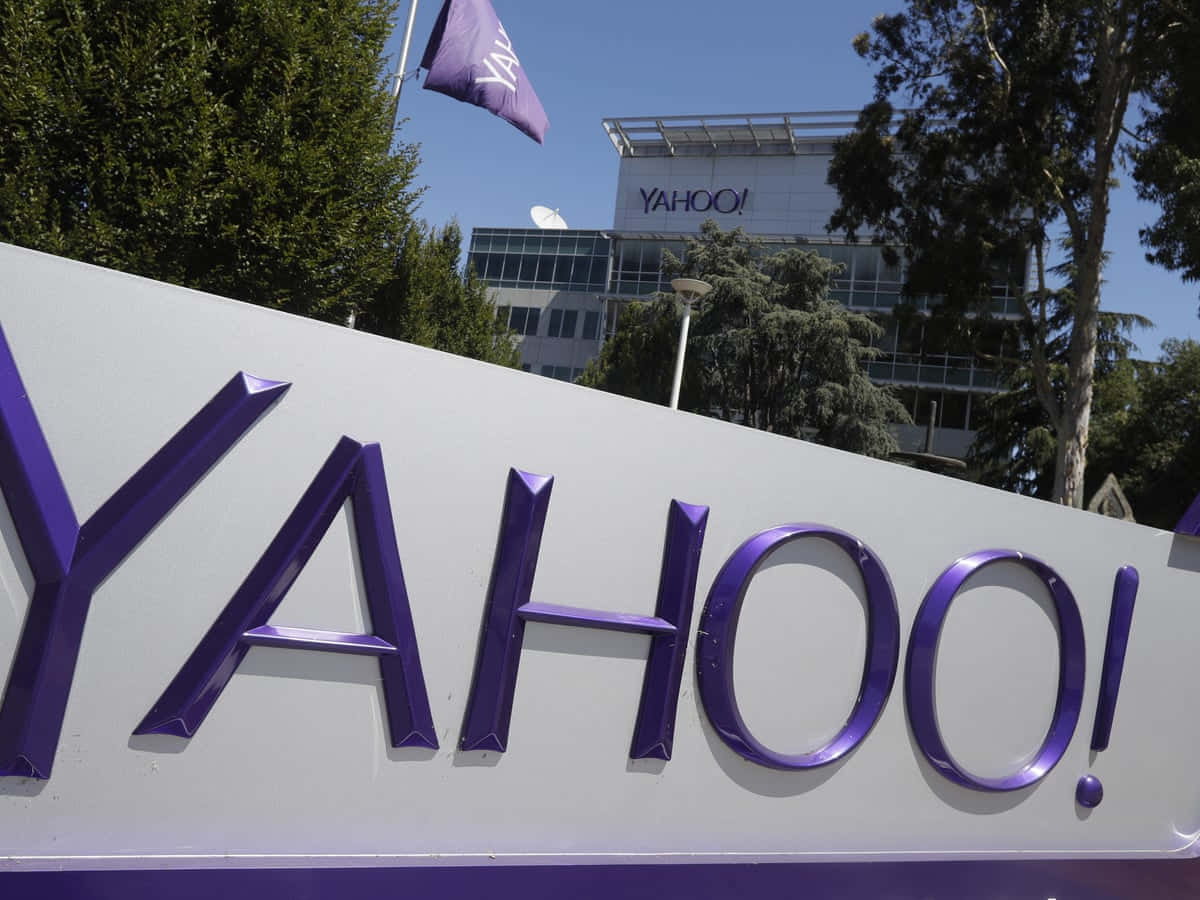 Yahoo! — 連結人們關注的重點 壁紙 — 一座現代玻璃建築前，有一個大型紫色Yahoo!標誌，周圍有樹木和晴朗的藍天。