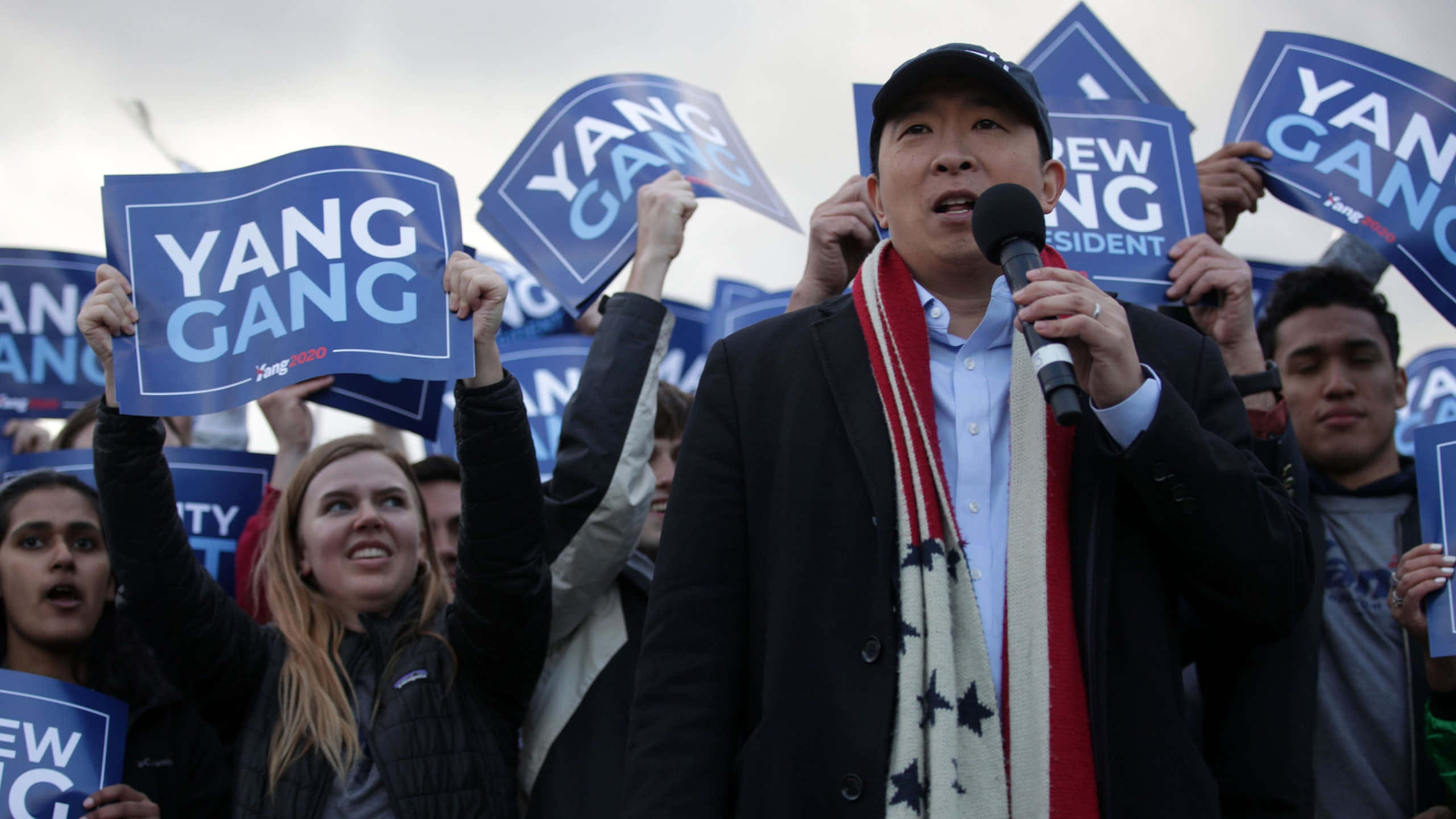 Andrew Yang 桌布 壁紙 — 楊安澤在戶外集會上對著麥克風講話，周圍是舉著藍色「YANG GANG」標語的支持者。