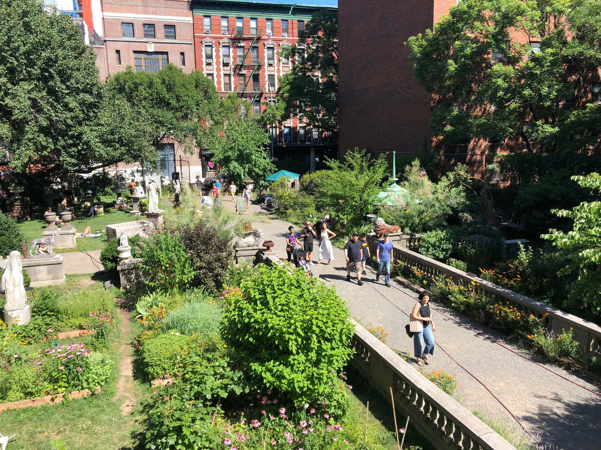 人們在陽光花園中行走，背景有雕像和磚砌建築。