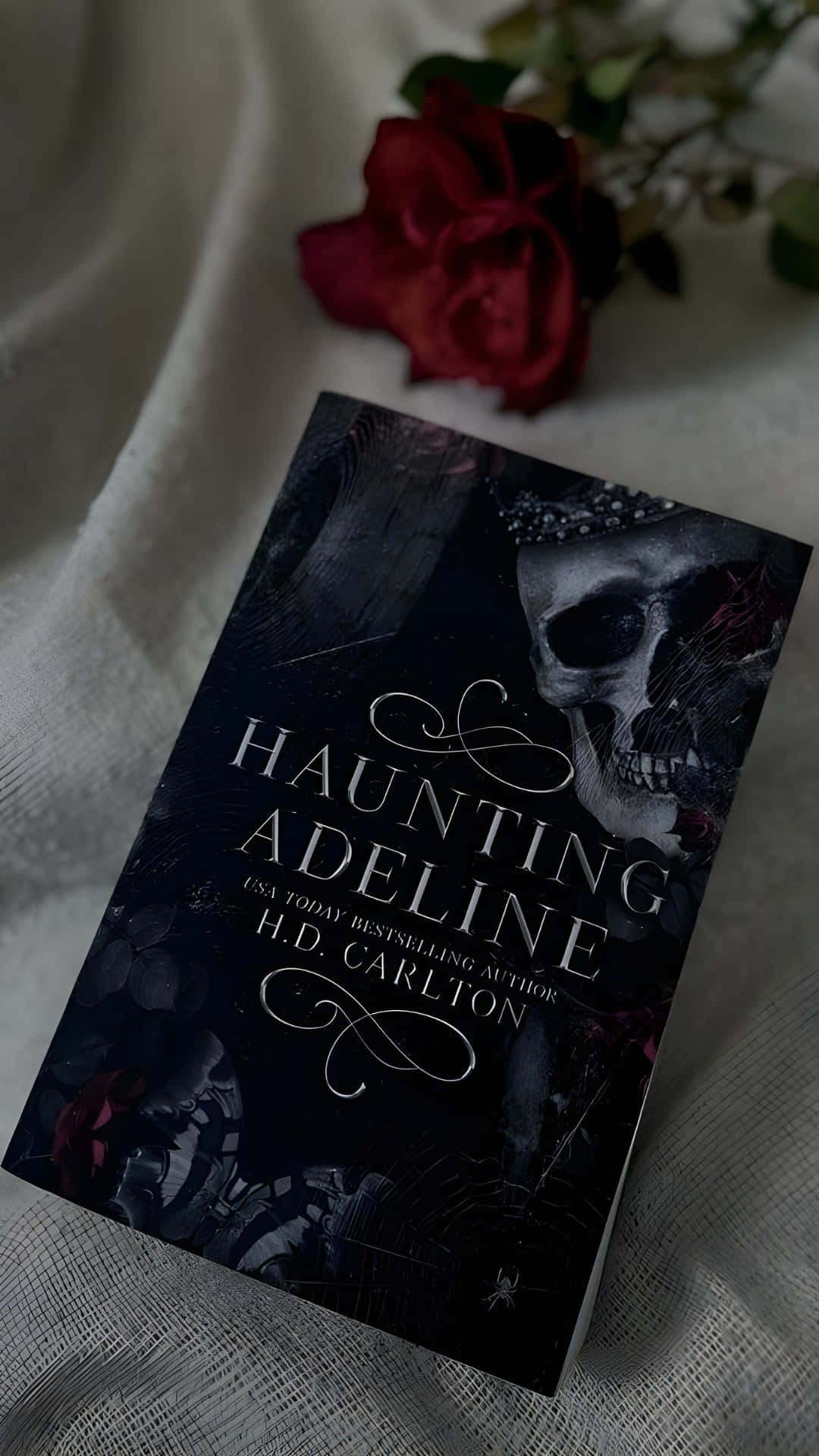  壁紙 — 一本名為《Haunting Adeline》的黑暗奇幻書本放在有紋理的白色布料上，旁邊有一朵深紅玫瑰。