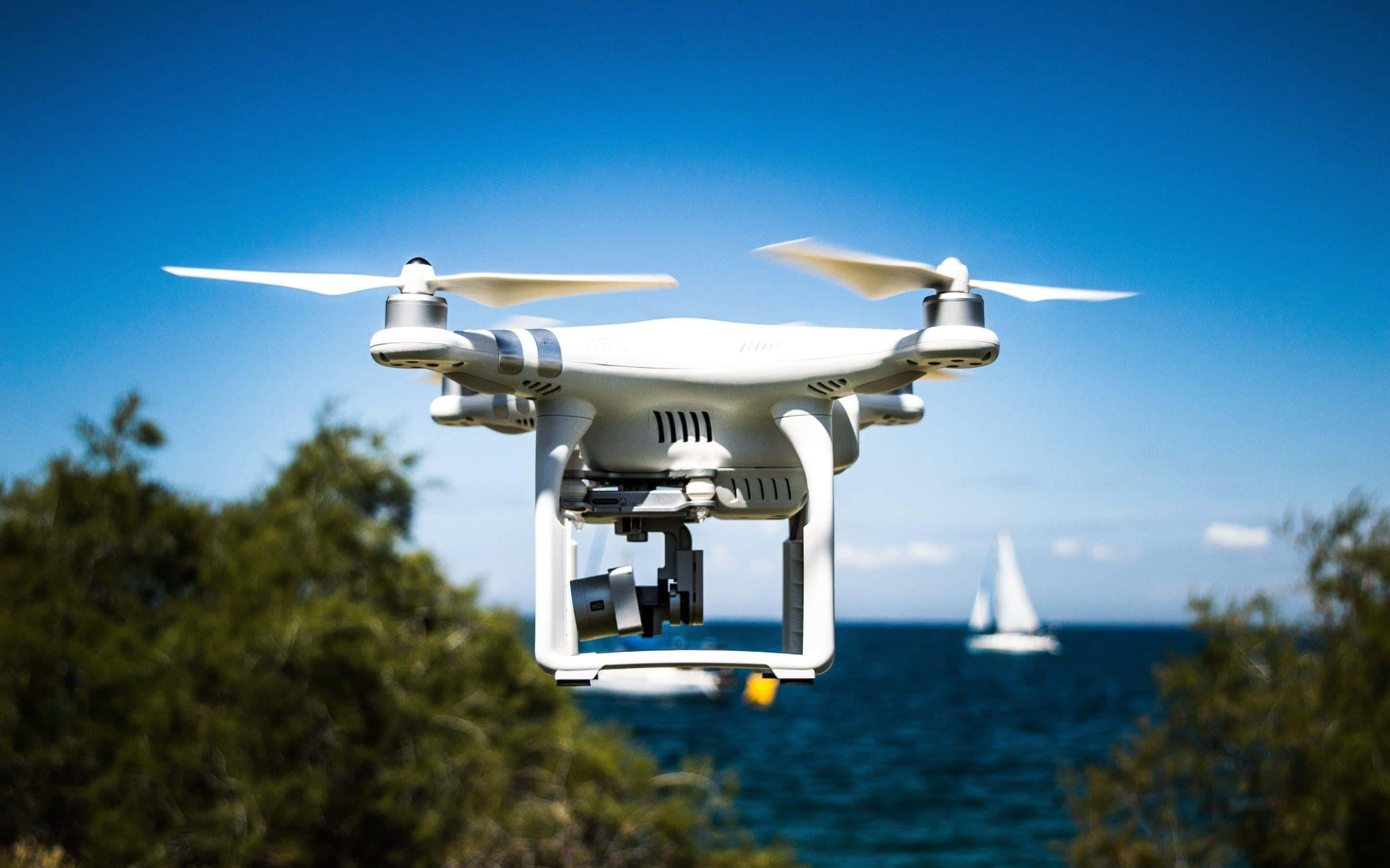 Drone Camera 桌布 壁紙 — 一架白色無人機在晴朗的天空下，盤旋於藍色海水上的帆船和綠色樹葉的海岸景觀之上。