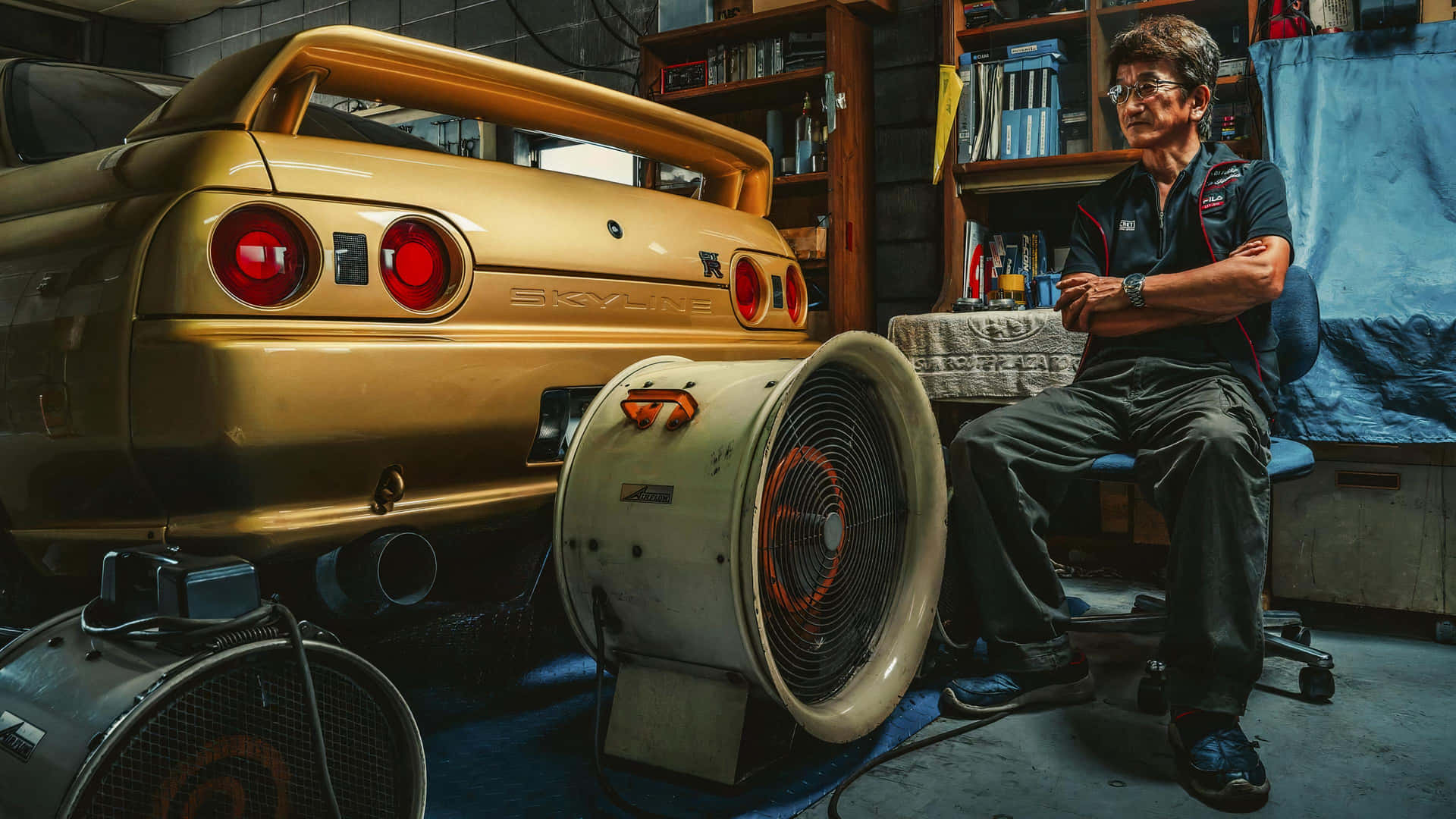  壁紙 — 一架金色日產Skyline R34停泊在雜亂的車房內，旁邊有一個大型工業風扇，一位身穿工作服的技師坐在椅子上沉思。