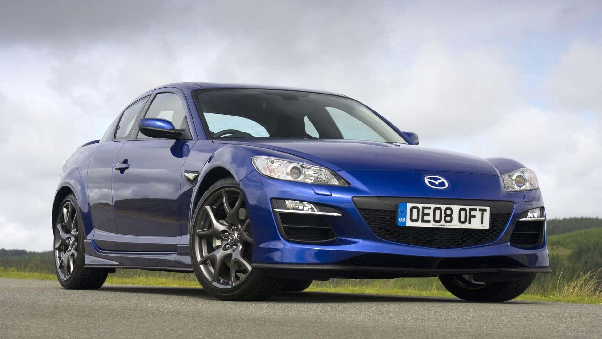 Mazda Rx-8 桌布 壁紙 — 一架光亮的藍色萬事得RX-8跑車停泊在瀝青路上，背景有草地山丘，天空多雲。