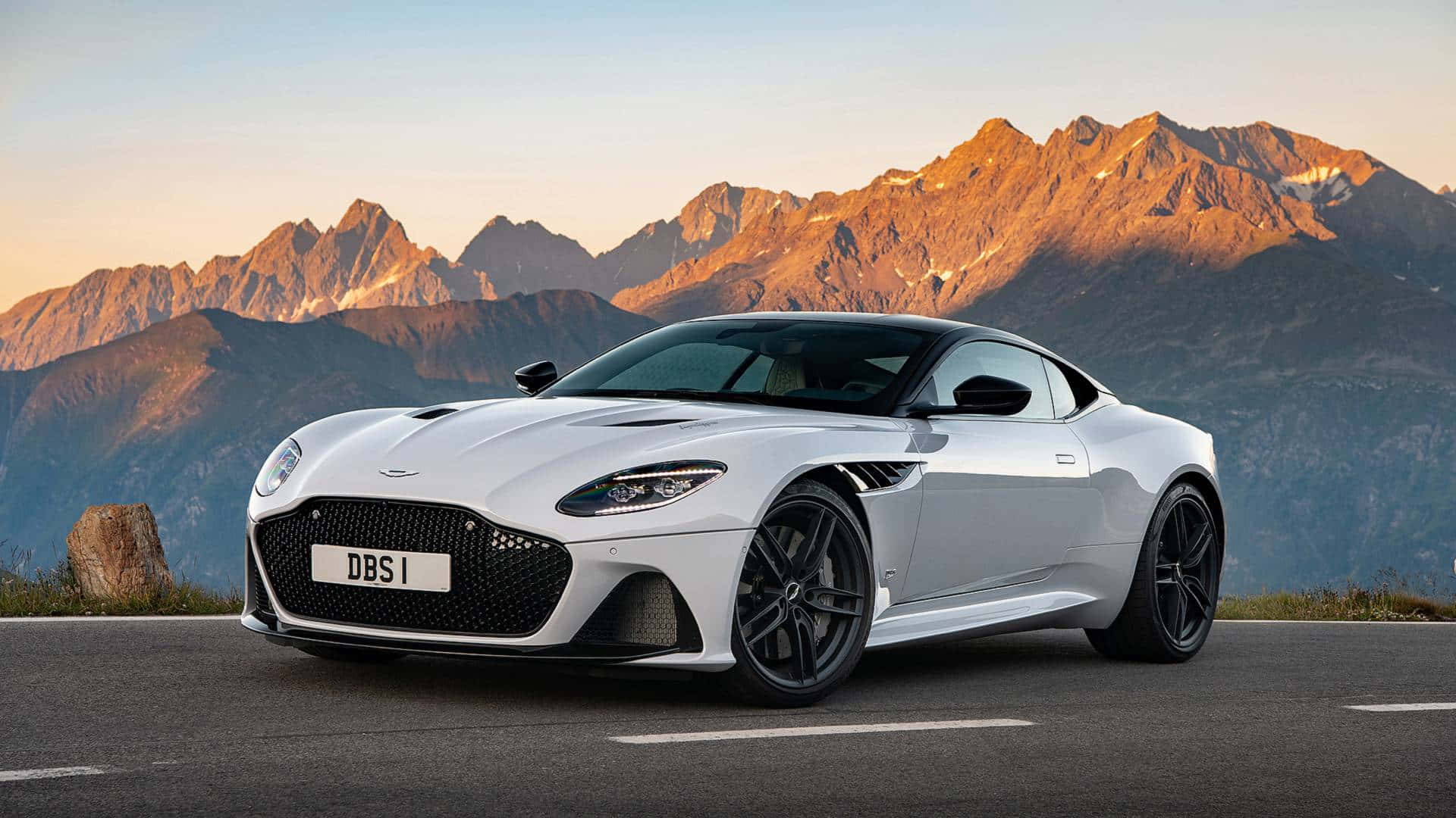 Aston Martin Dbs Superleggera 桌布 壁紙 — 一架流線型白色Aston Martin DBS I跑車停泊在山路上，背景是日落時分金色的山峰。