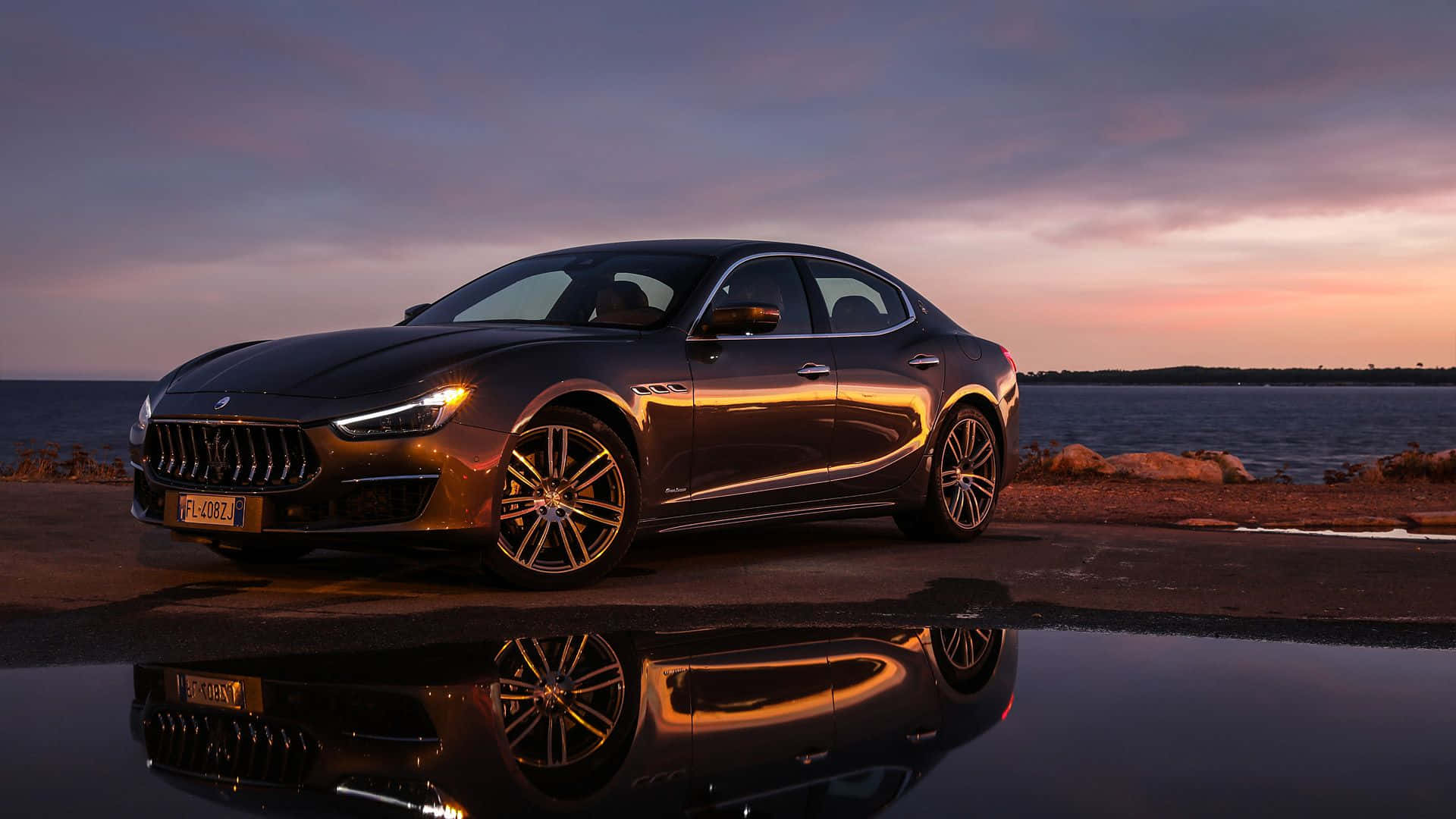 Maserati Ghibli 桌布 壁紙 — 一架流線型黑色瑪莎拉蒂房車在黃昏閃耀，倒影映照在海邊道路旁的水坑中。