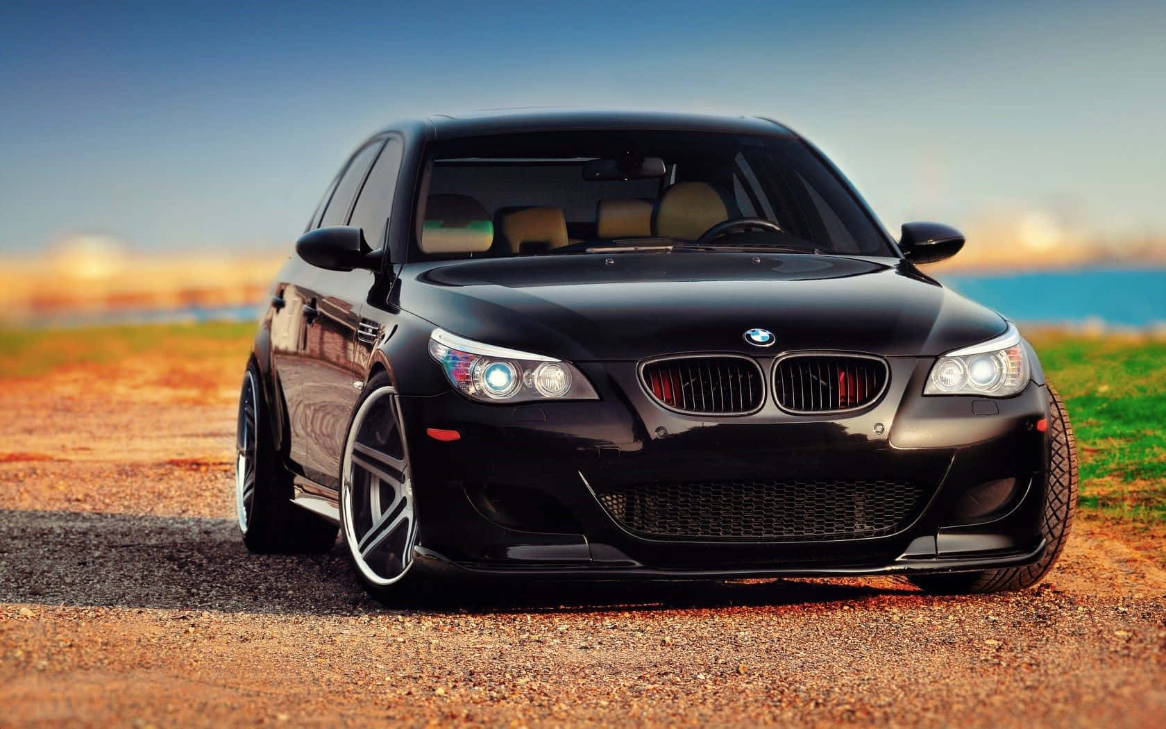 Bmw M5 桌布 壁紙 — 一架亮黑色BMW M5運動轎車停泊在碎石路上，背景是模糊的海岸景觀。