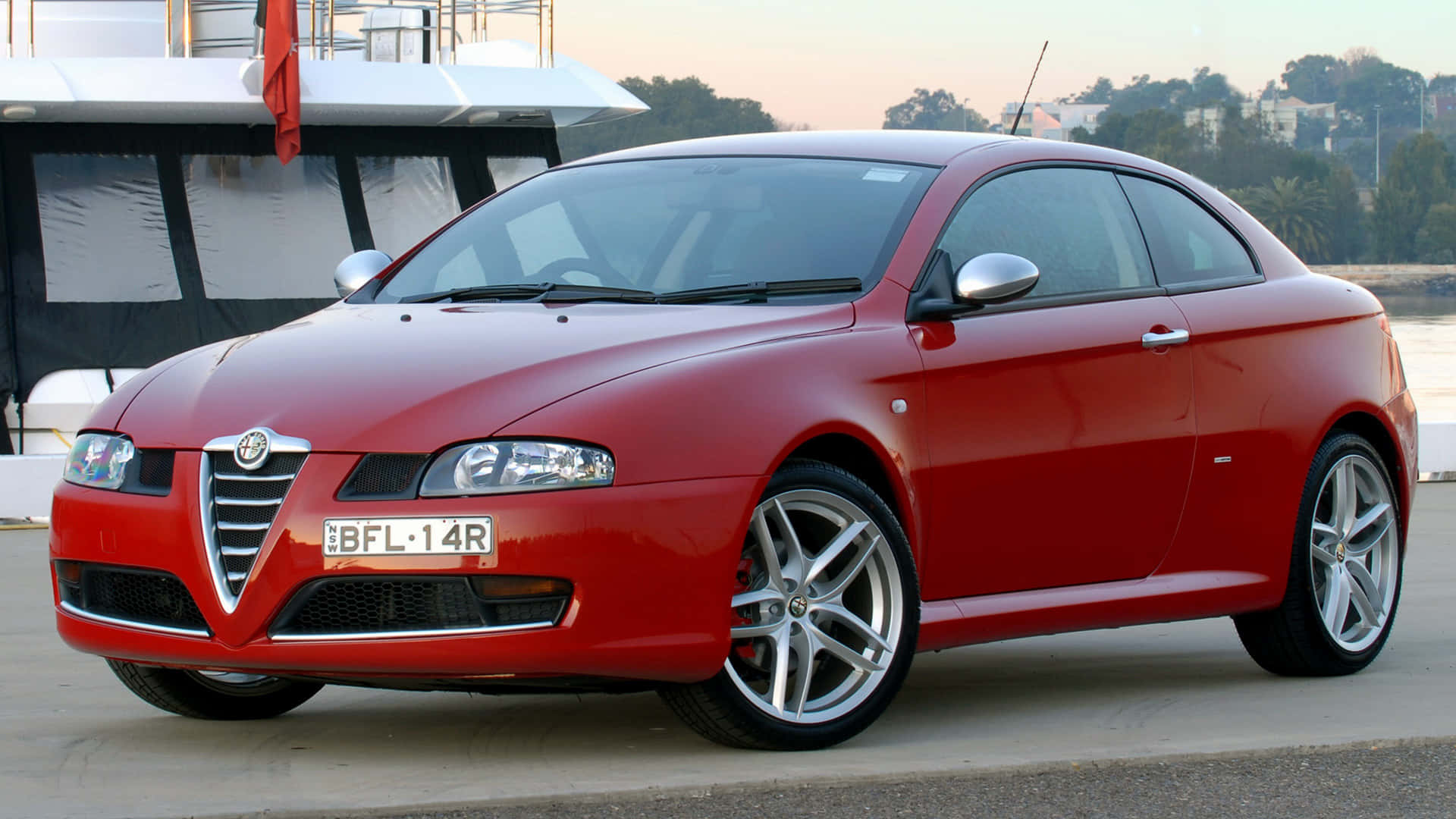 Alfa Romeo Gt 桌布 壁紙 — 一架亮紅色愛快羅密歐轎跑車停泊在遊艇旁，夕陽西下，溫暖的環境光線反射在其流暢的曲線上。