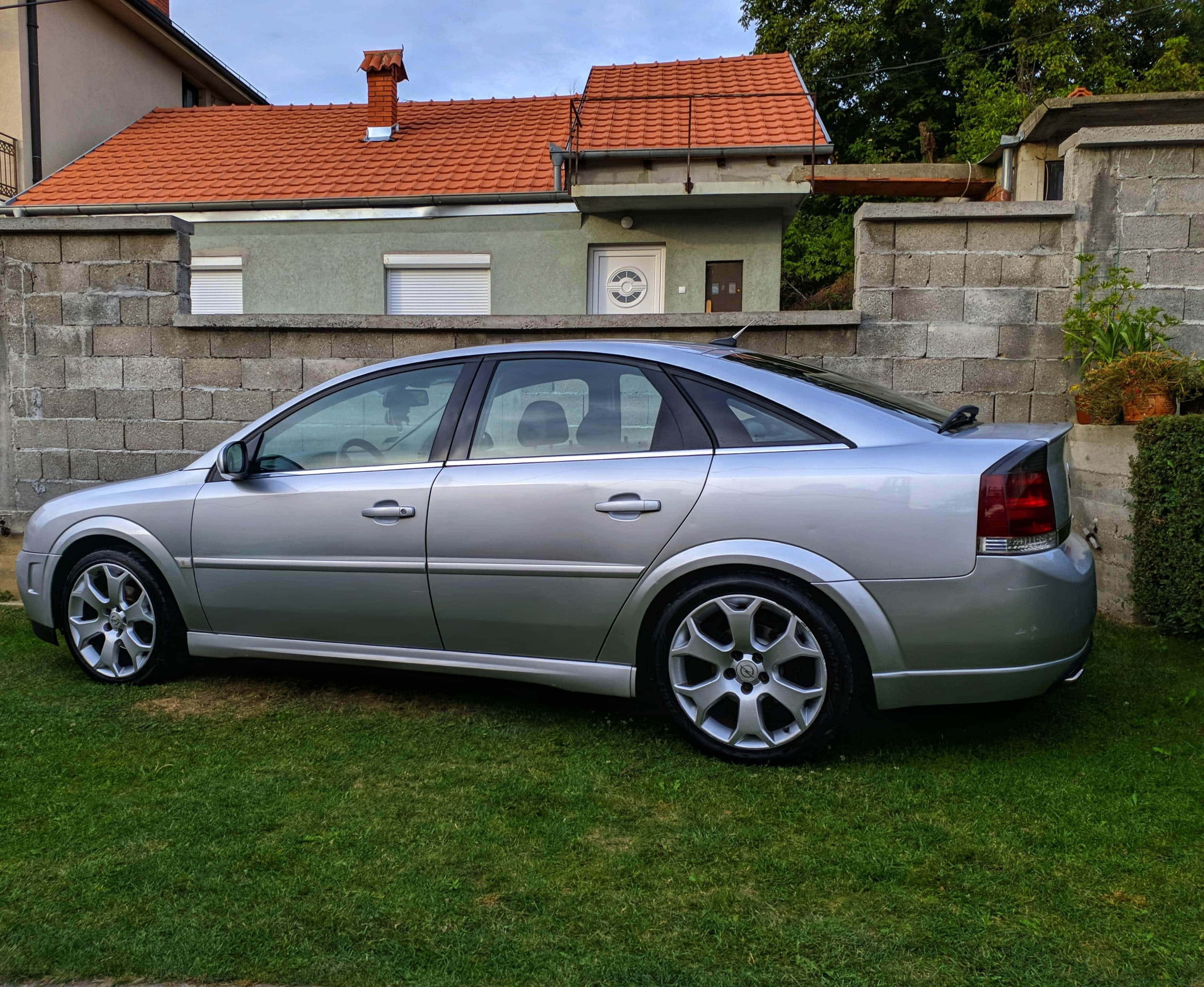 Opel Vectra 桌布 壁紙 — 一架銀色房車停泊在石牆和瓦頂屋前的綠色草地上，天空多雲。