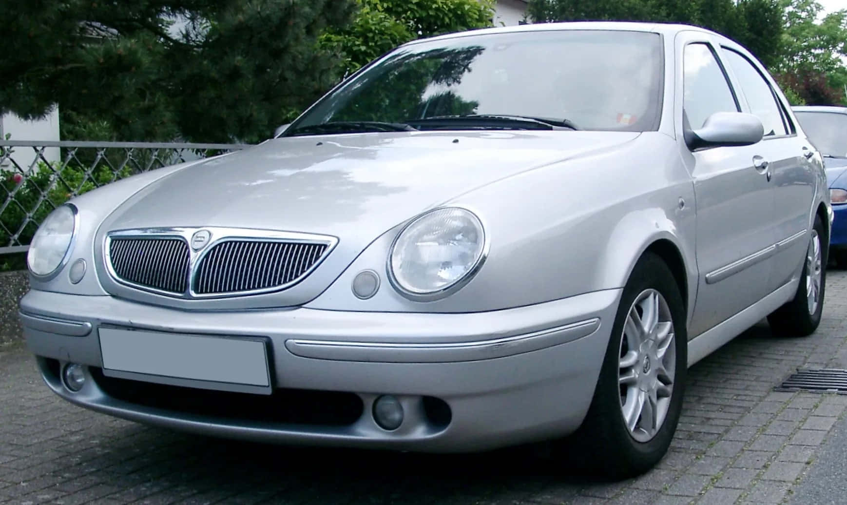  壁紙 — 一架銀色 Lancia 房車停泊在鋪砌的車道上，背景有綠樹和圍欄。