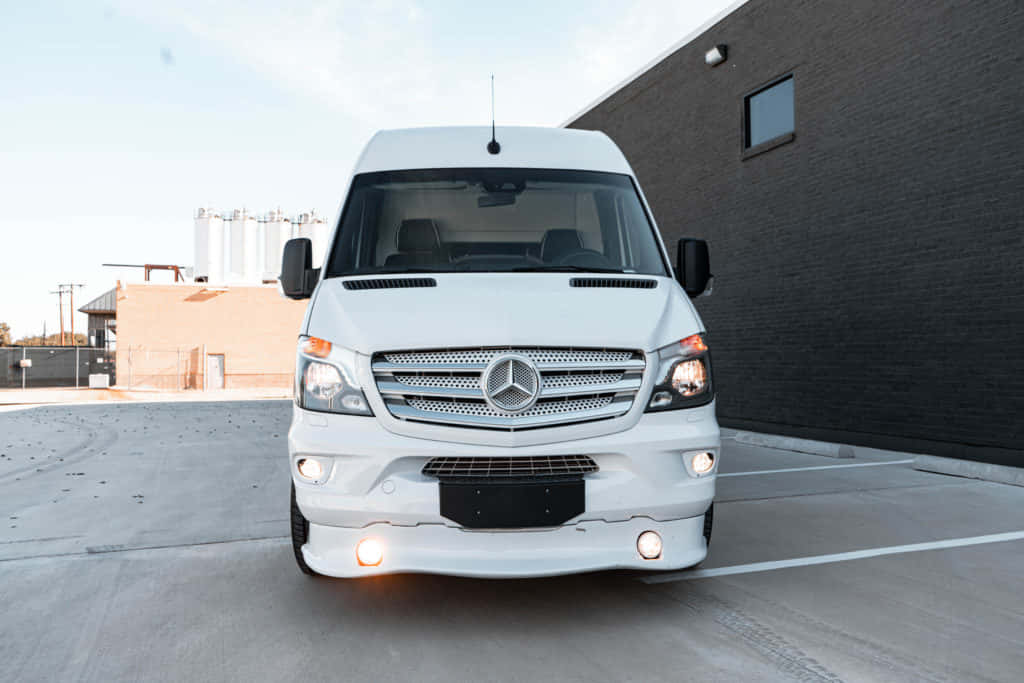 Mercedes Benz Sprinter 桌布 壁紙 — 一架純白色的 Mercedes-Benz Sprinter 客貨車停泊在混凝土停車場，旁邊是深色磚砌建築，天空晴朗。