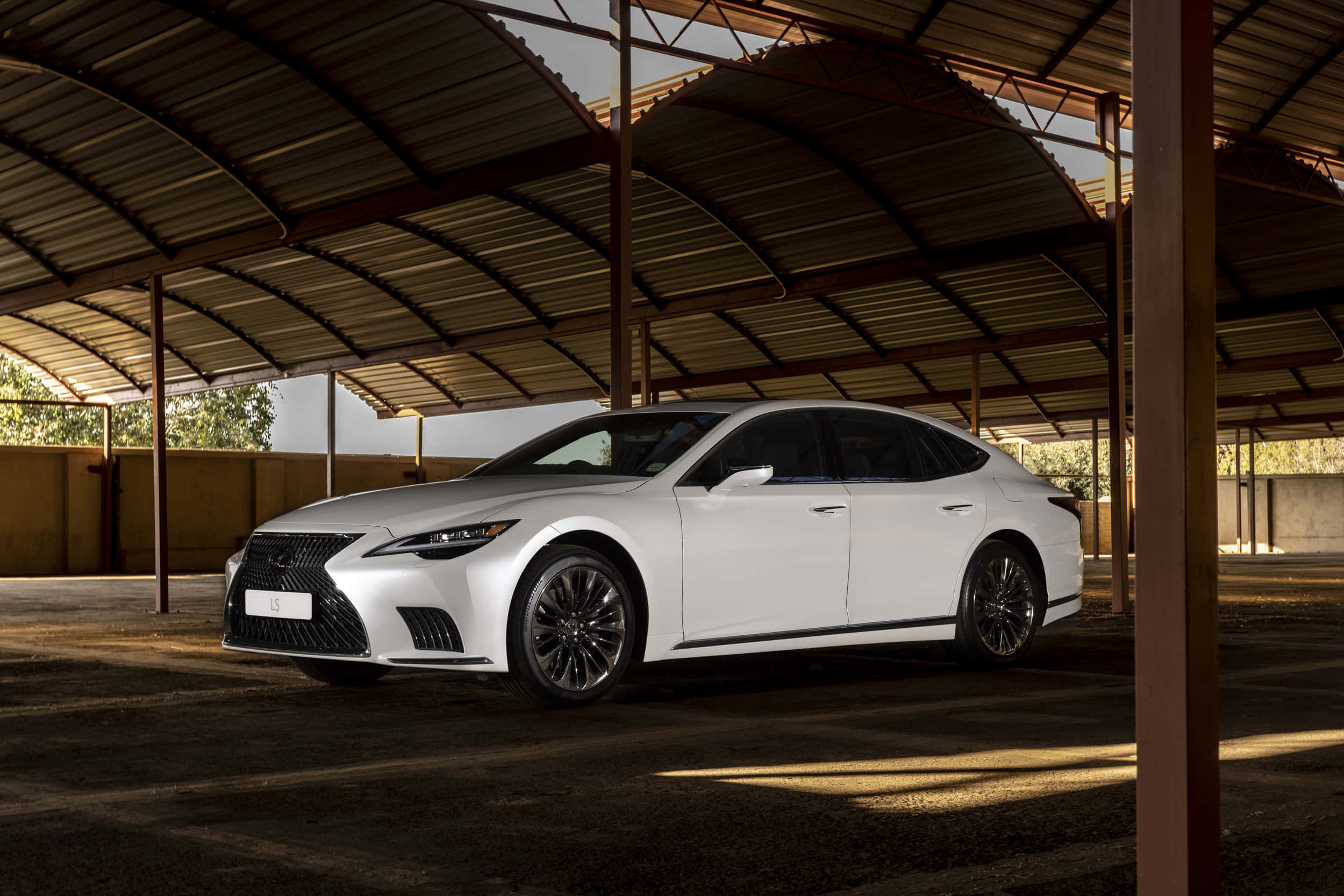 Lexus Ls 500 桌布 壁紙 — 一架時尚的白色Lexus房車停泊在金屬屋頂下，陽光在塵土飛揚的地面上投下長長的影子。