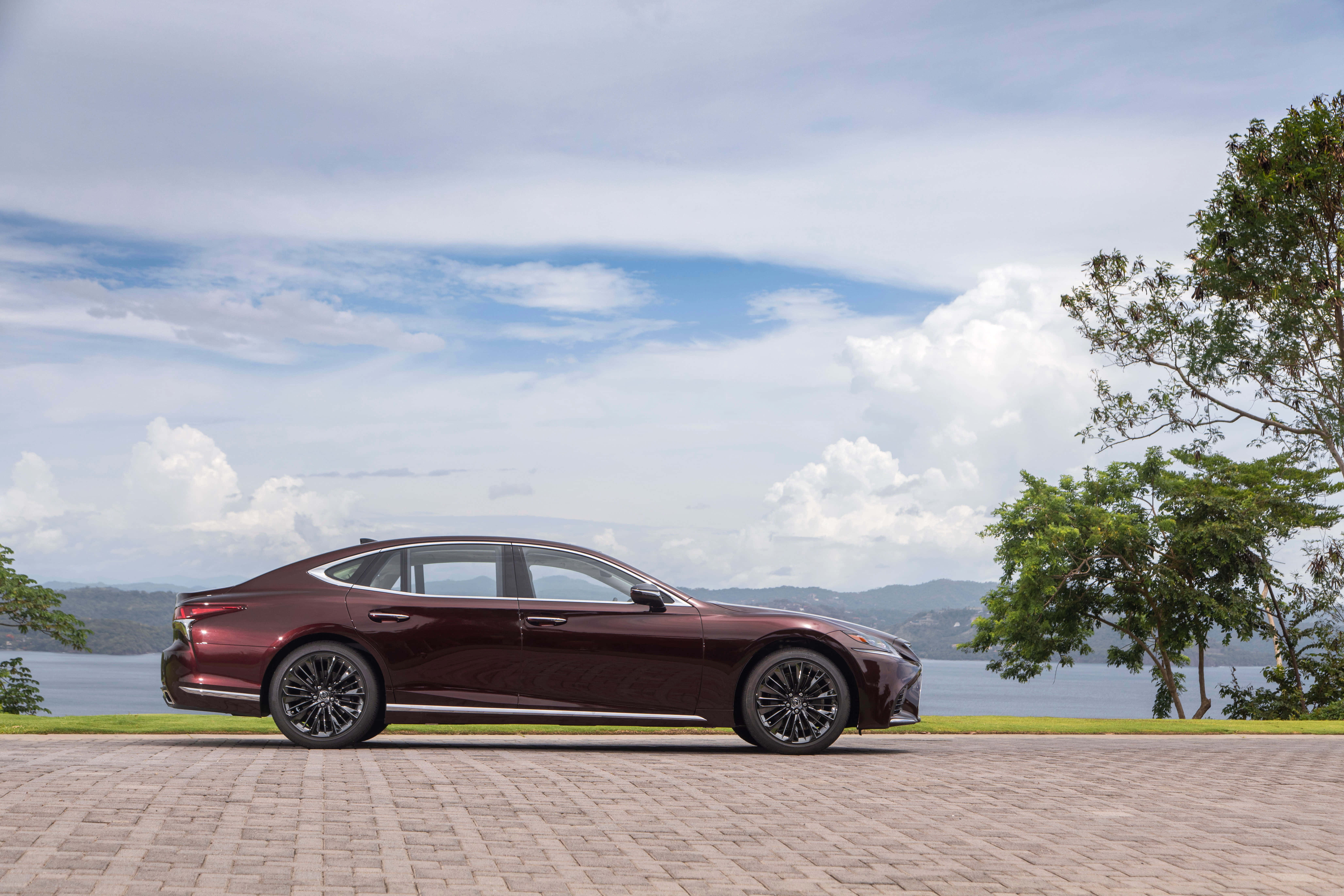 Lexus Ls 500 桌布 壁紙 — 一架時尚的酒紅色轎車停在磚砌車道上，旁邊是湖泊，遠處有山丘，頭頂有蓬鬆的雲朵。
