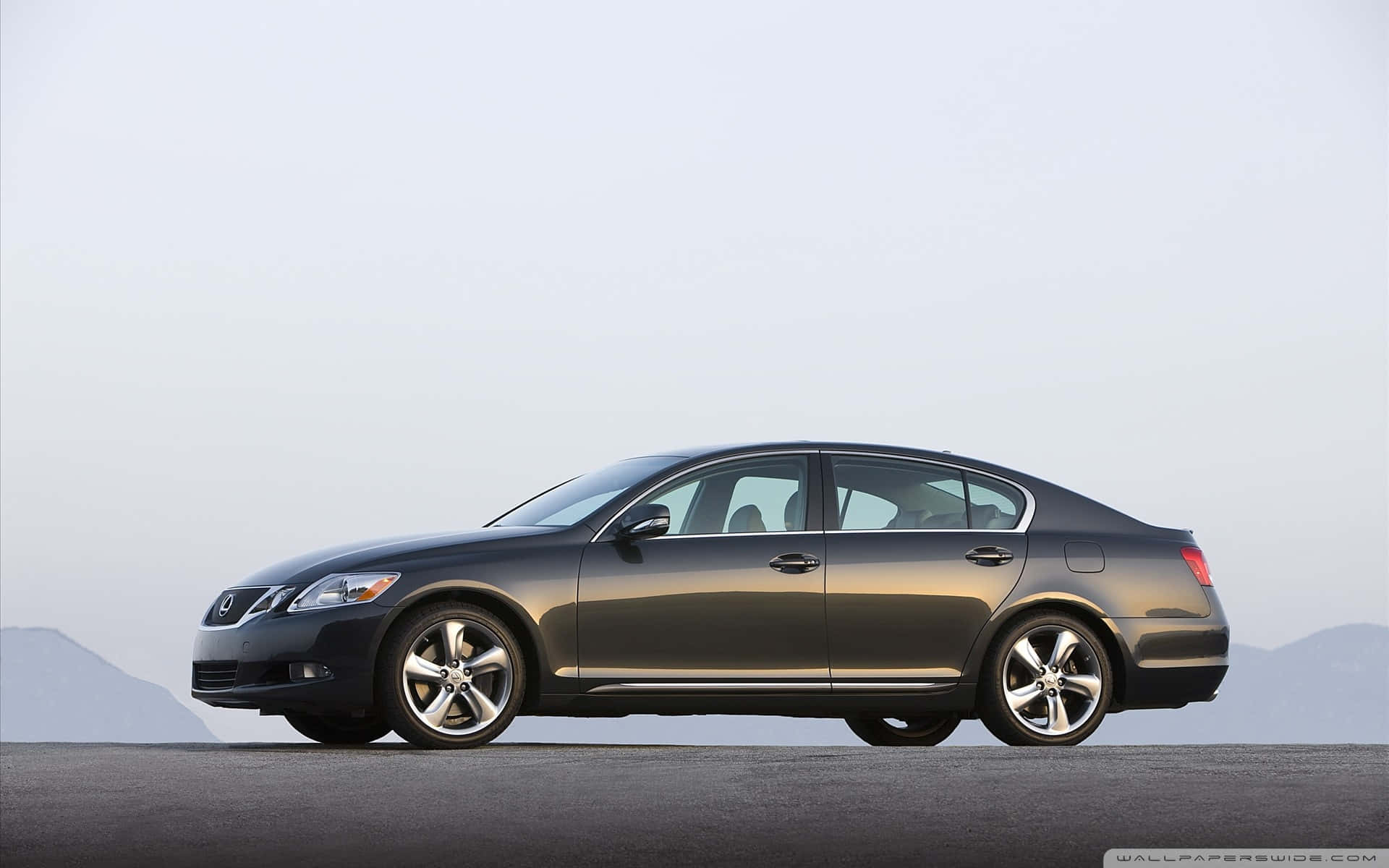 Lexus Gs 桌布 壁紙 — 一架時尚的深灰色轎車停泊在鋪砌的道路上，遠處有山脈，天空朦朧。