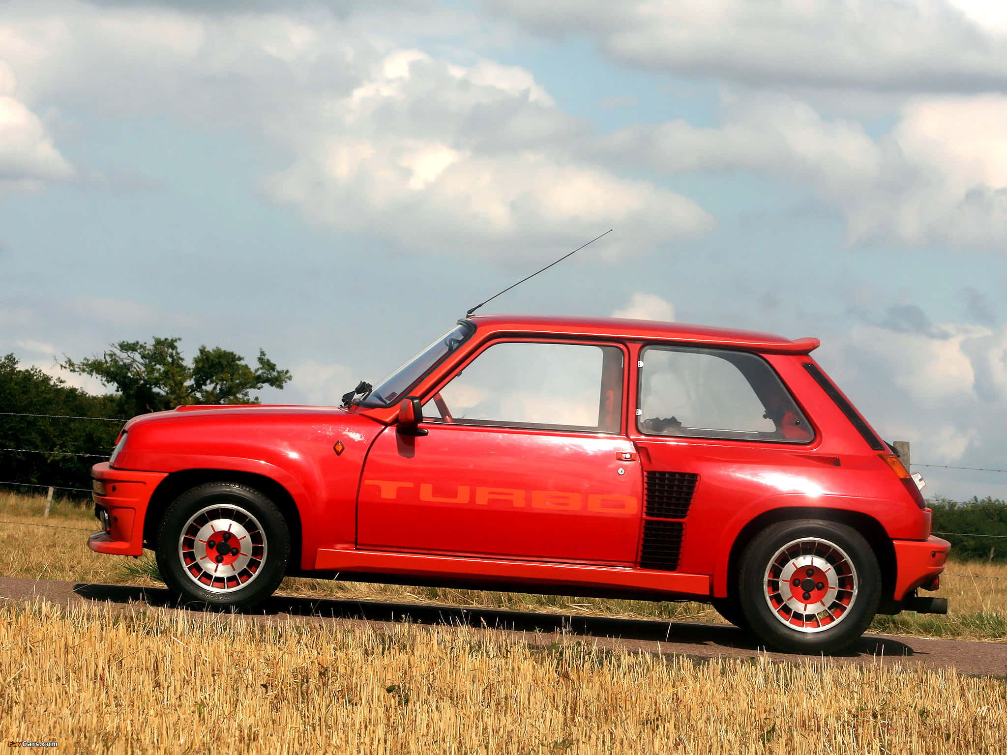 Renault 5 Turbo 桌布 壁紙 — 一架鮮紅色嘅雷諾5 Turbo跑車停喺鄉村道路旁邊，金色草叢之上，天空多雲。