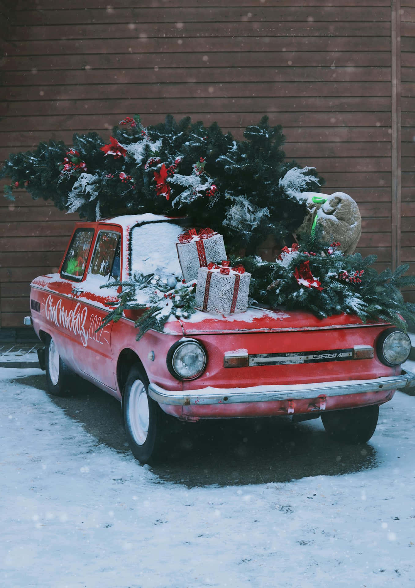  壁紙 — 一架覆蓋著雪的復古紅色汽車，載著裝飾好的聖誕樹和禮物，背景是木牆。