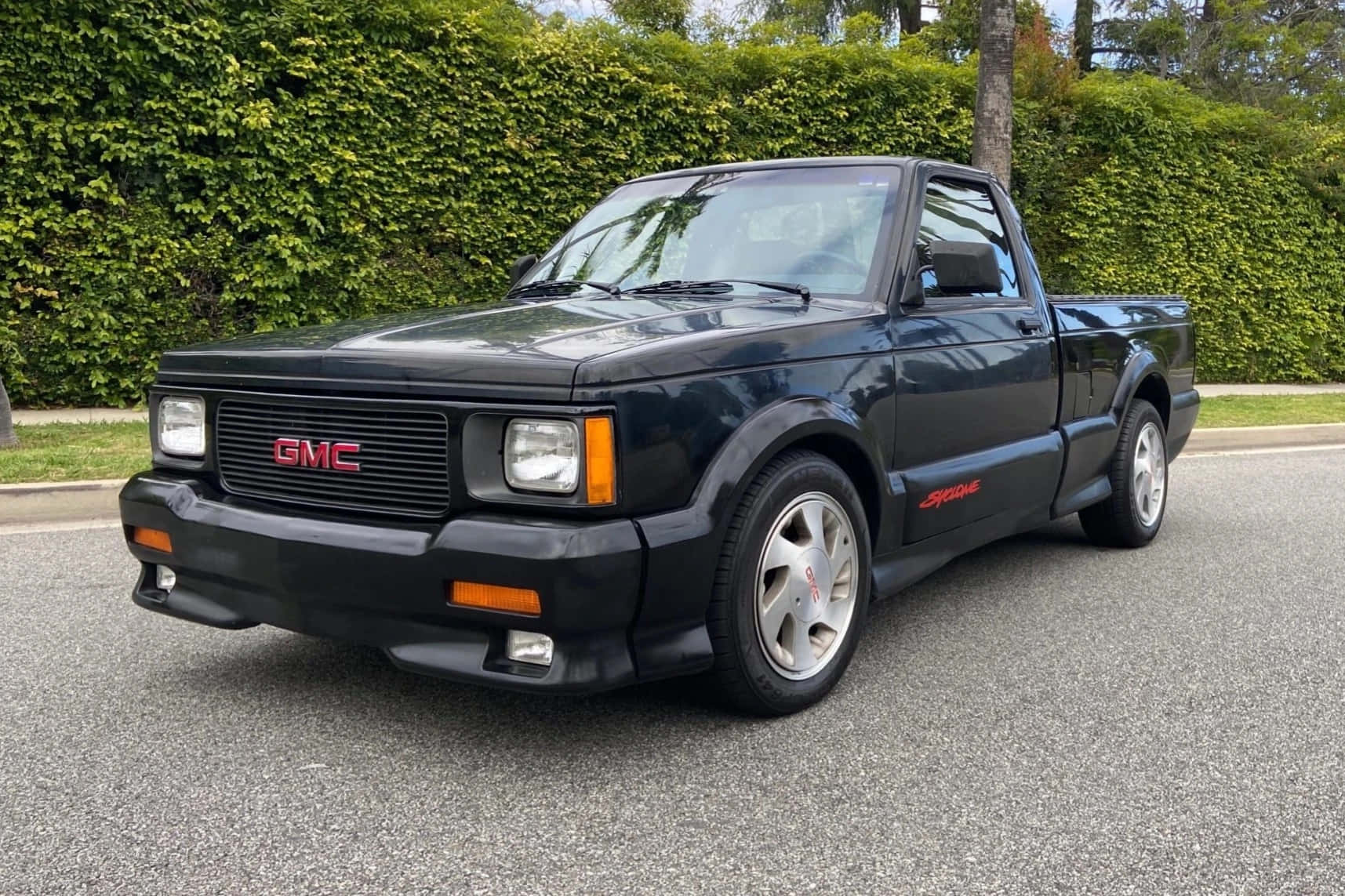 Gmc Syclone 桌布 壁紙 — 一架型格黑色GMC Syclone皮卡車停在瀝青路上，背景是茂密的綠色樹籬。
