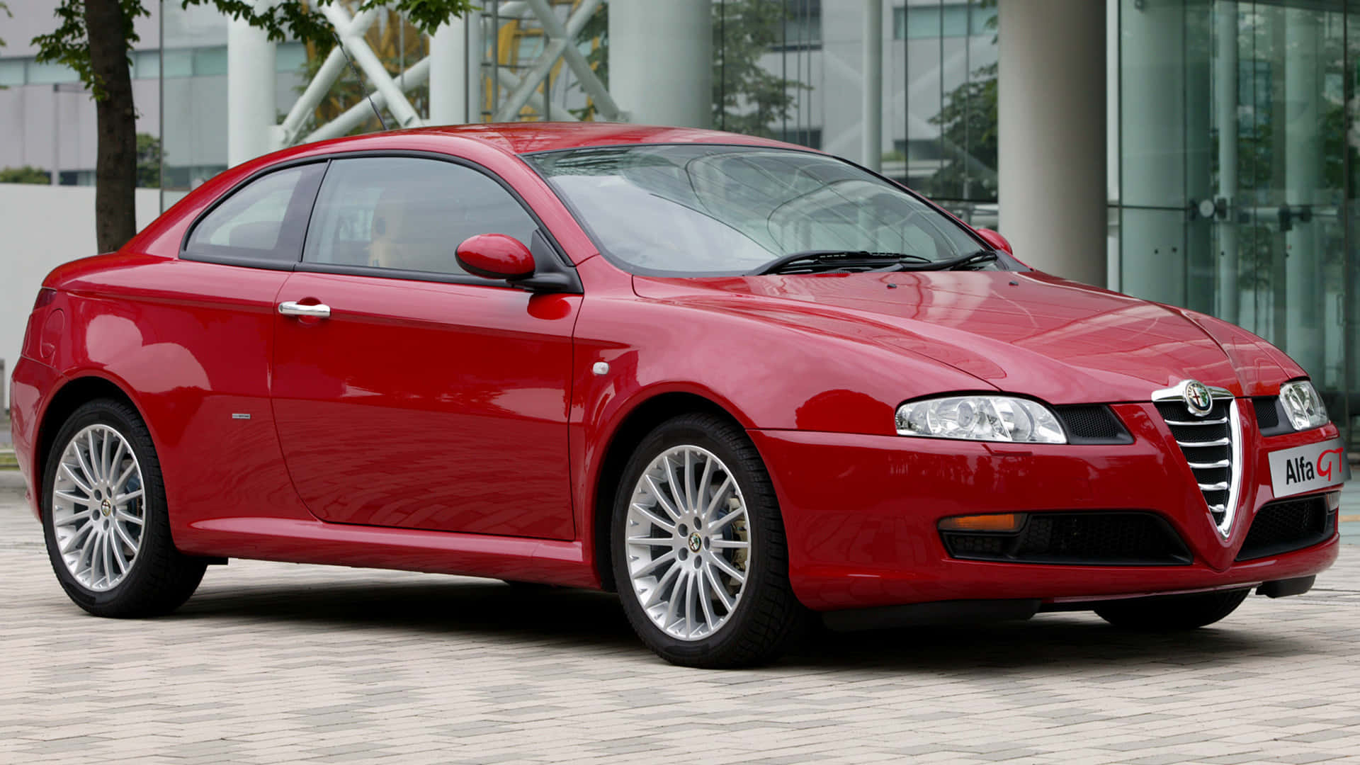 Alfa Romeo Gt 桌布 壁紙 — 一架亮紅色嘅Alfa Romeo GT轎跑車停泊喺鋪砌嘅廣場上，背景係現代玻璃建築。