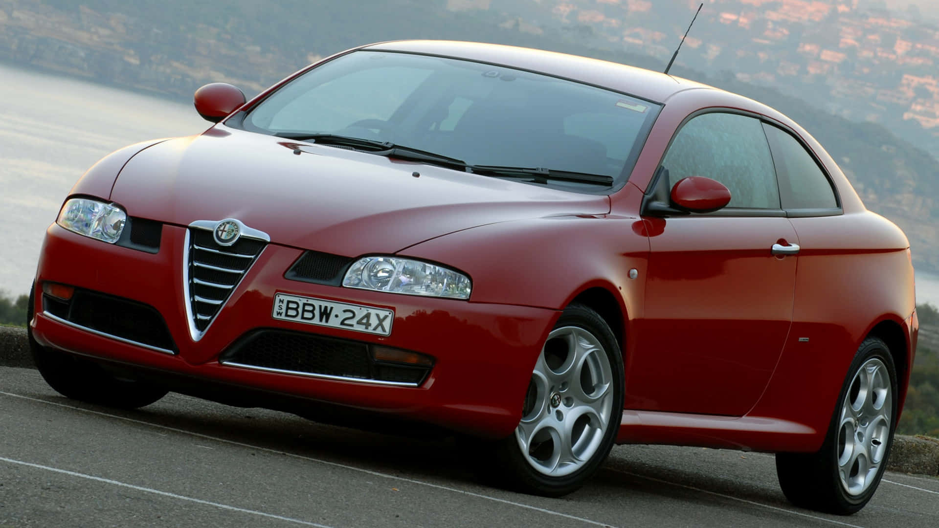 Alfa Romeo Gt 桌布 壁紙 — 一架亮紅色嘅愛快羅密歐轎跑車停泊喺沿海公路，背景係模糊嘅城市景觀。