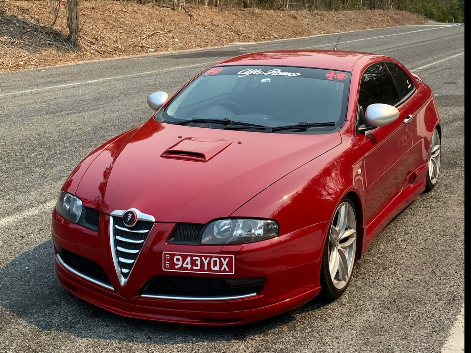 Alfa Romeo Gt 桌布 壁紙 — 一架亮麗的紅色愛快羅密歐跑車停泊在陽光照耀的瀝青路上，背景有乾草和樹木。
