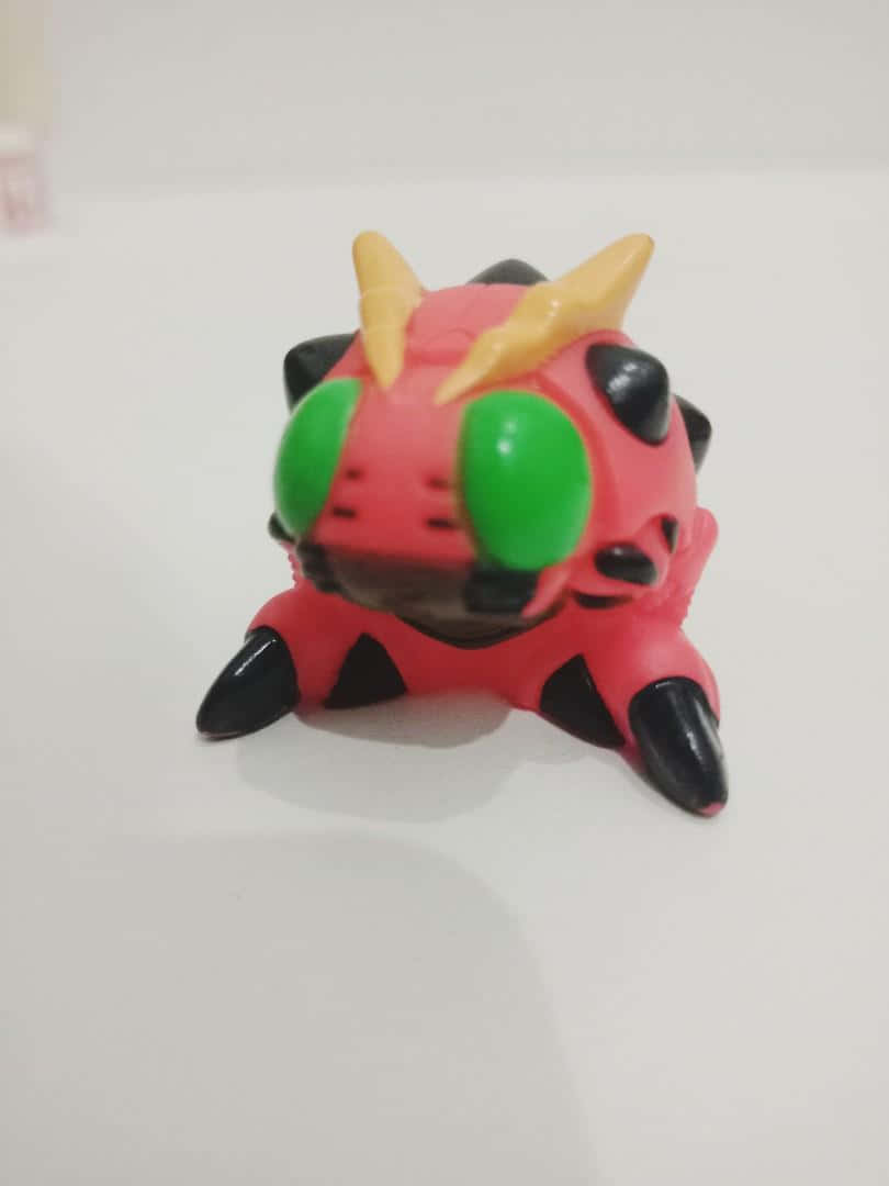 Digimon Tentomon 桌布 壁紙 — 一個細小嘅紅色塑膠玩具怪獸，有綠色眼睛、黑色爪同黃色角，坐喺一個純白色嘅表面上。