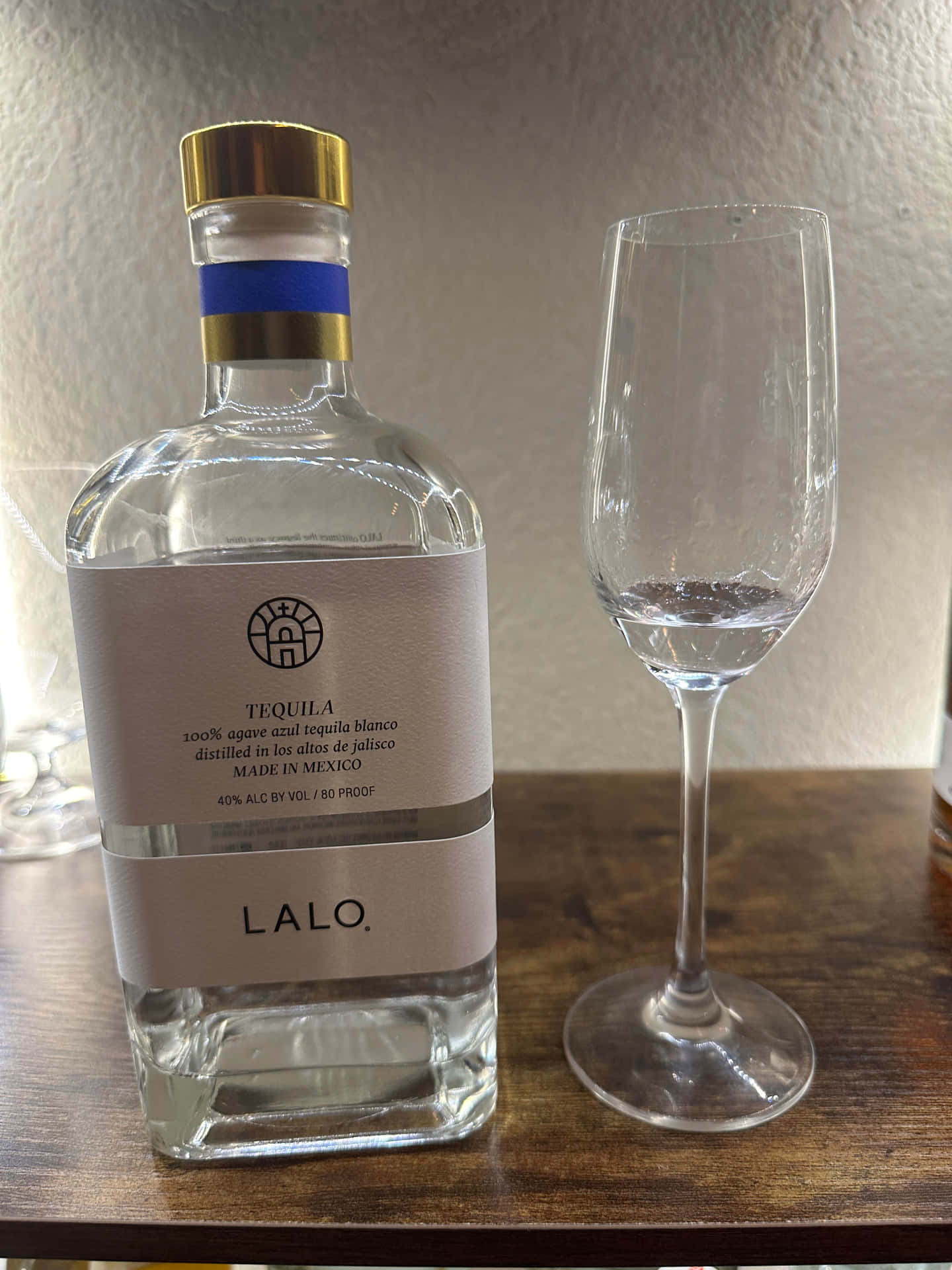  壁紙 — 一個透明嘅Lalo龍舌蘭酒樽，金色樽蓋，放喺木枱上一個空嘅酒杯旁邊。