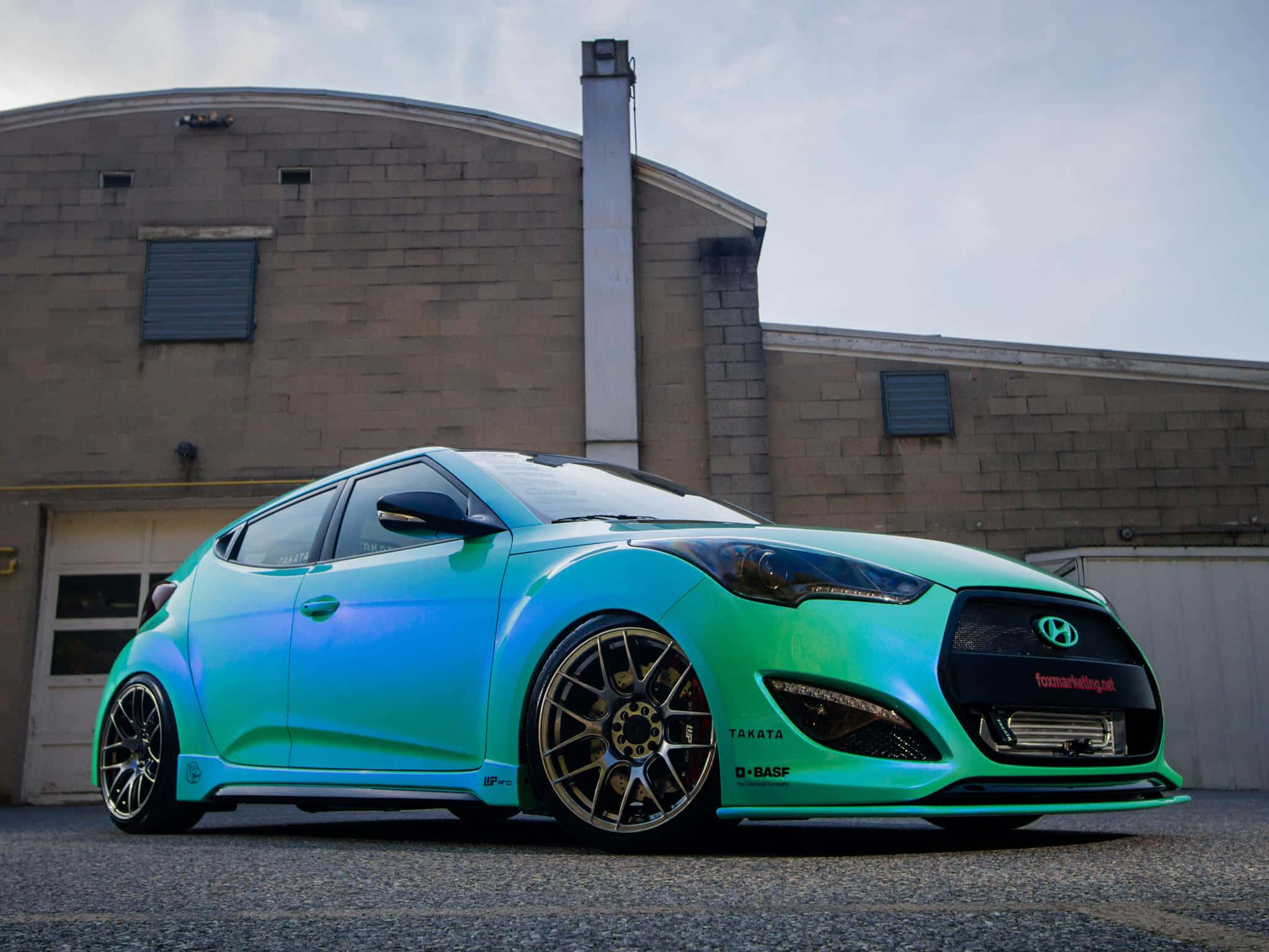 Hyundai Veloster 桌布 壁紙 — 一架充滿活力的漸變青綠色和藍色改裝現代Veloster停在一棟磚造工業建築前。