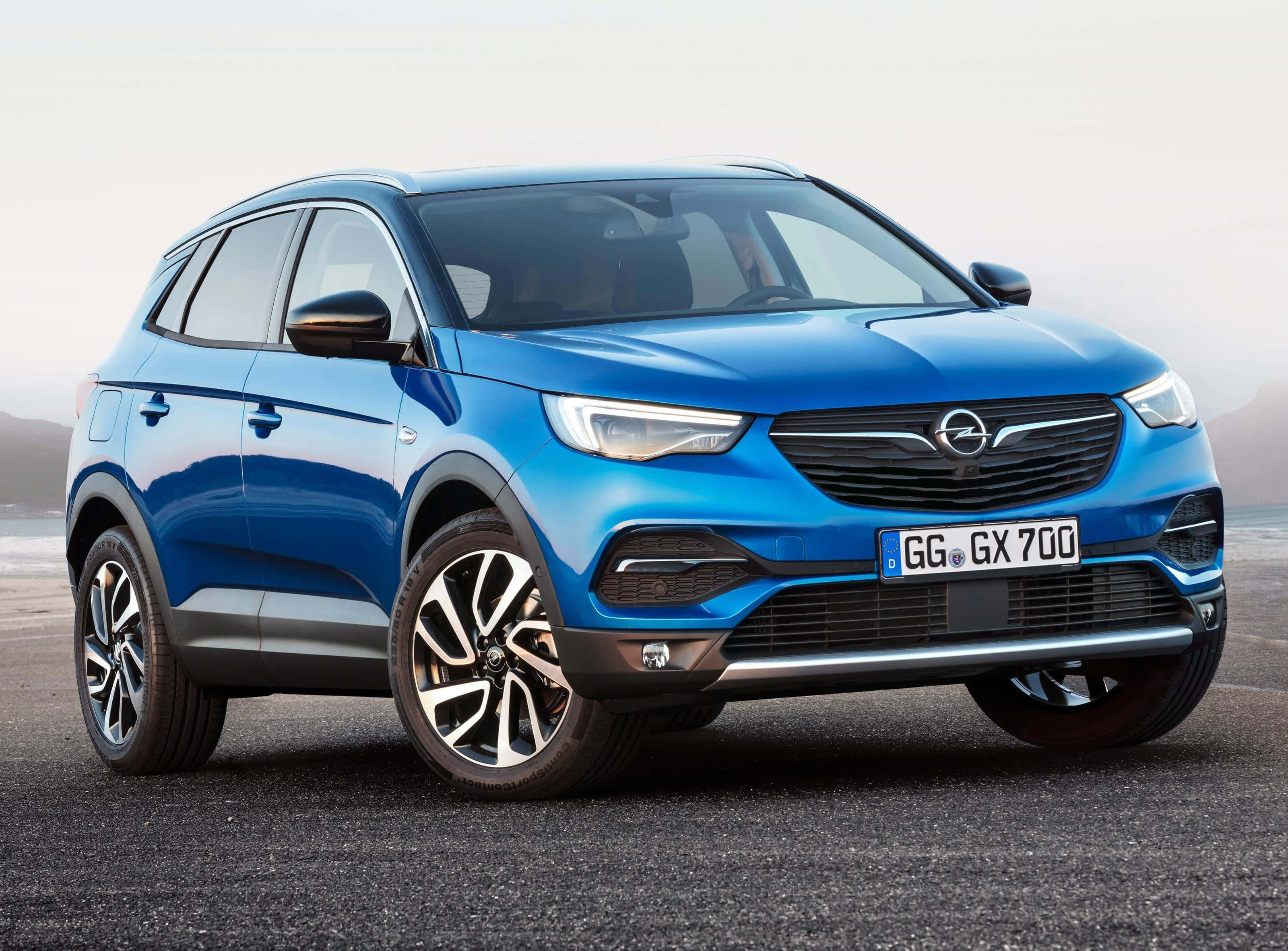 Opel Grandland X 桌布 壁紙 — 一架鮮藍色歐寶SUV停在瀝青路上，背景有朦朧的山脈，在柔和日光下。