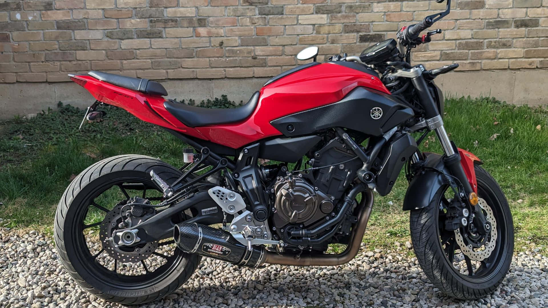  壁紙 — 一架搶眼的紅黑色Yamaha電單車停泊在碎石上，背景係磚牆同綠色草地。