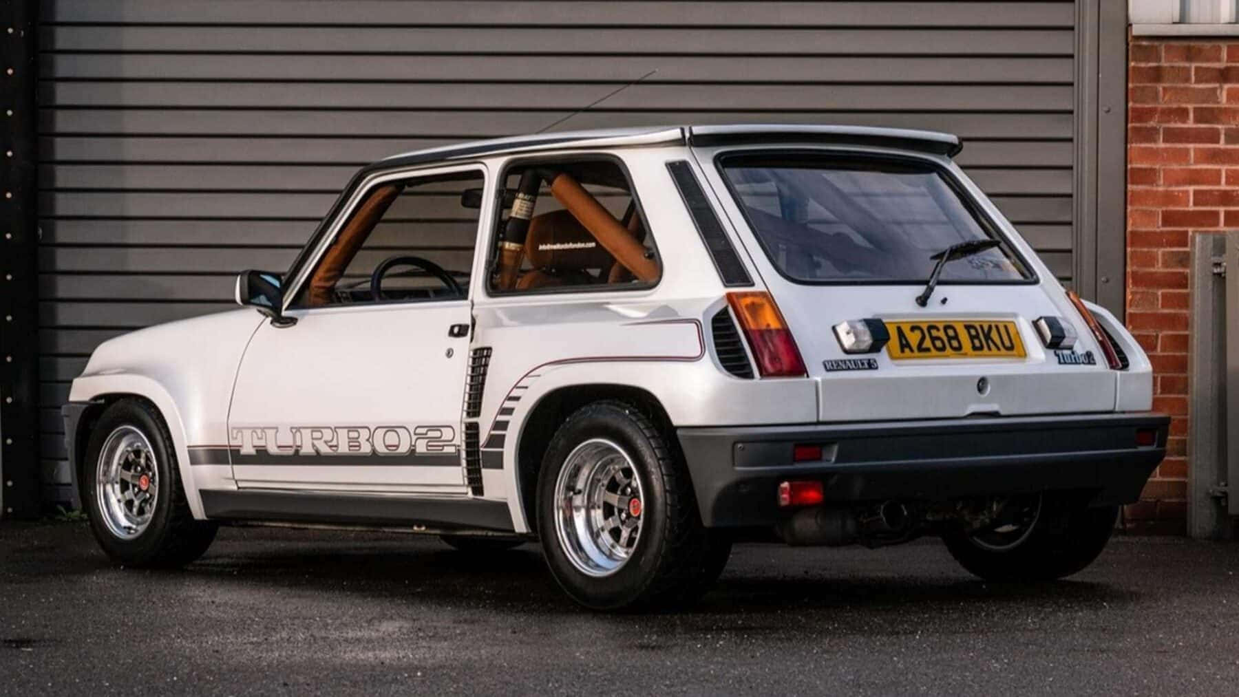 Renault 5 Turbo 桌布 壁紙 — 一輛白色雷諾5 Turbo 2跑車停泊在磚牆和金屬車庫門旁邊。