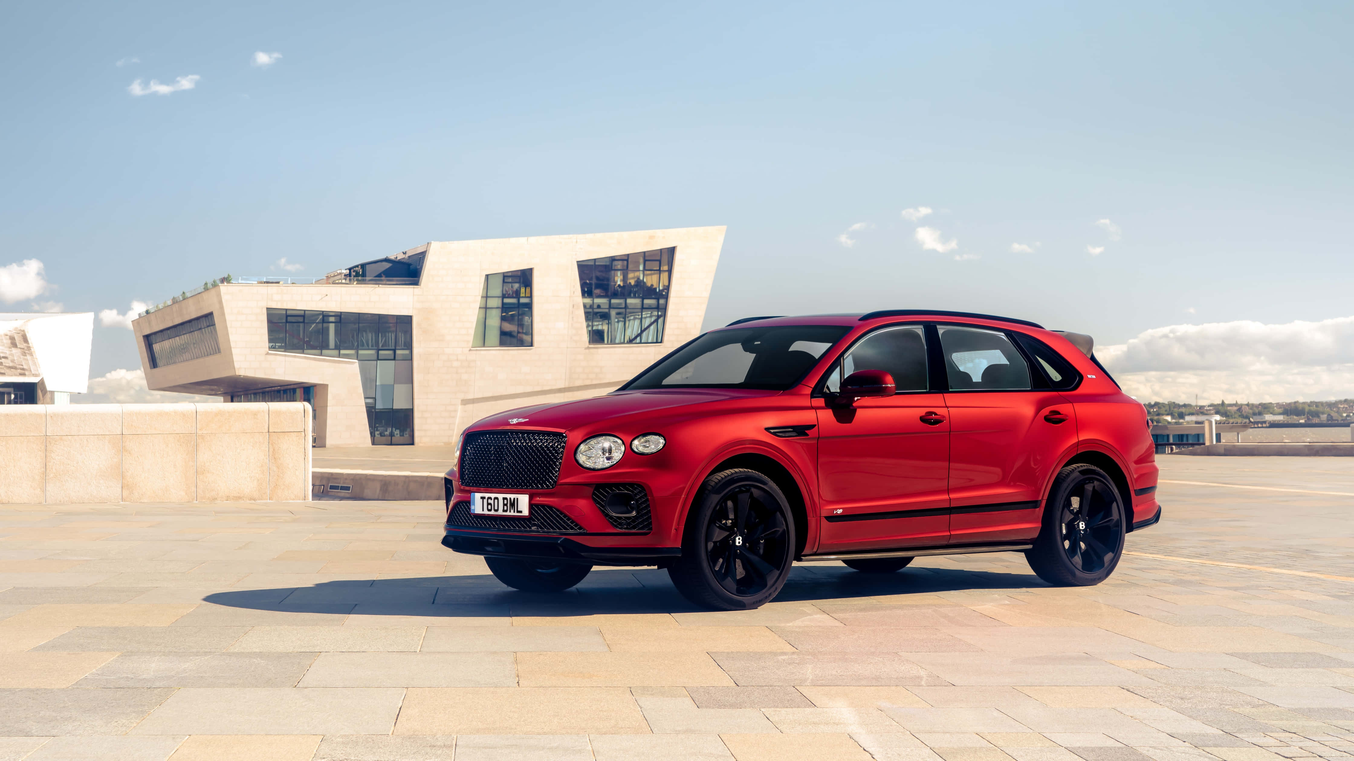 Bentley Bentayga 桌布 壁紙 — 一輛搶眼的紅色賓利Bentayga SUV停泊在石頭廣場上，旁邊是現代米色建築，天空清澈湛藍。