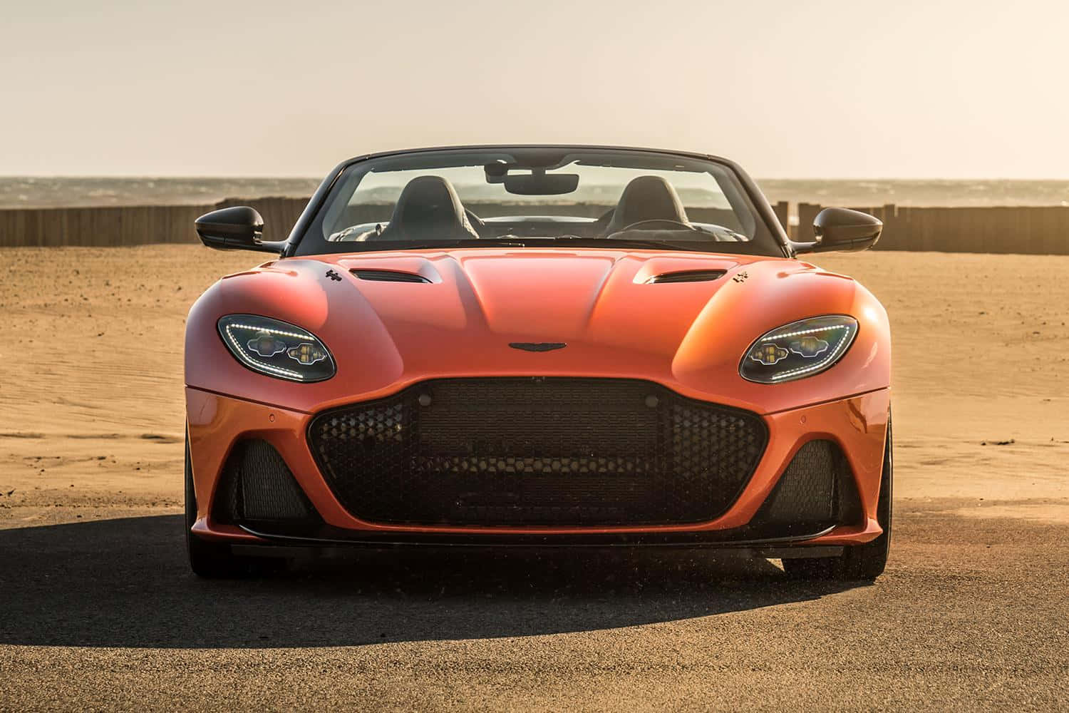 Aston Martin Dbs Superleggera 桌布 壁紙 — 一輛充滿活力的橙色 Aston Martin DBS Superleggera 開篷車停泊在陽光明媚的沙漠道路上，有木柵欄和遙遠的地平線。