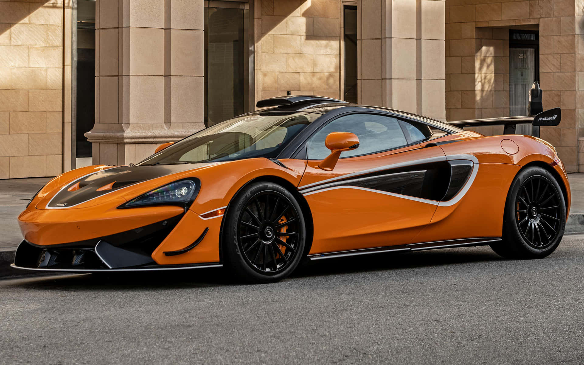 Mclaren 620r 桌布 壁紙 — 一輛充滿活力的橙色McLaren跑車，帶有黑色飾邊，在陽光明媚的日子停在一棟石頭建築前。