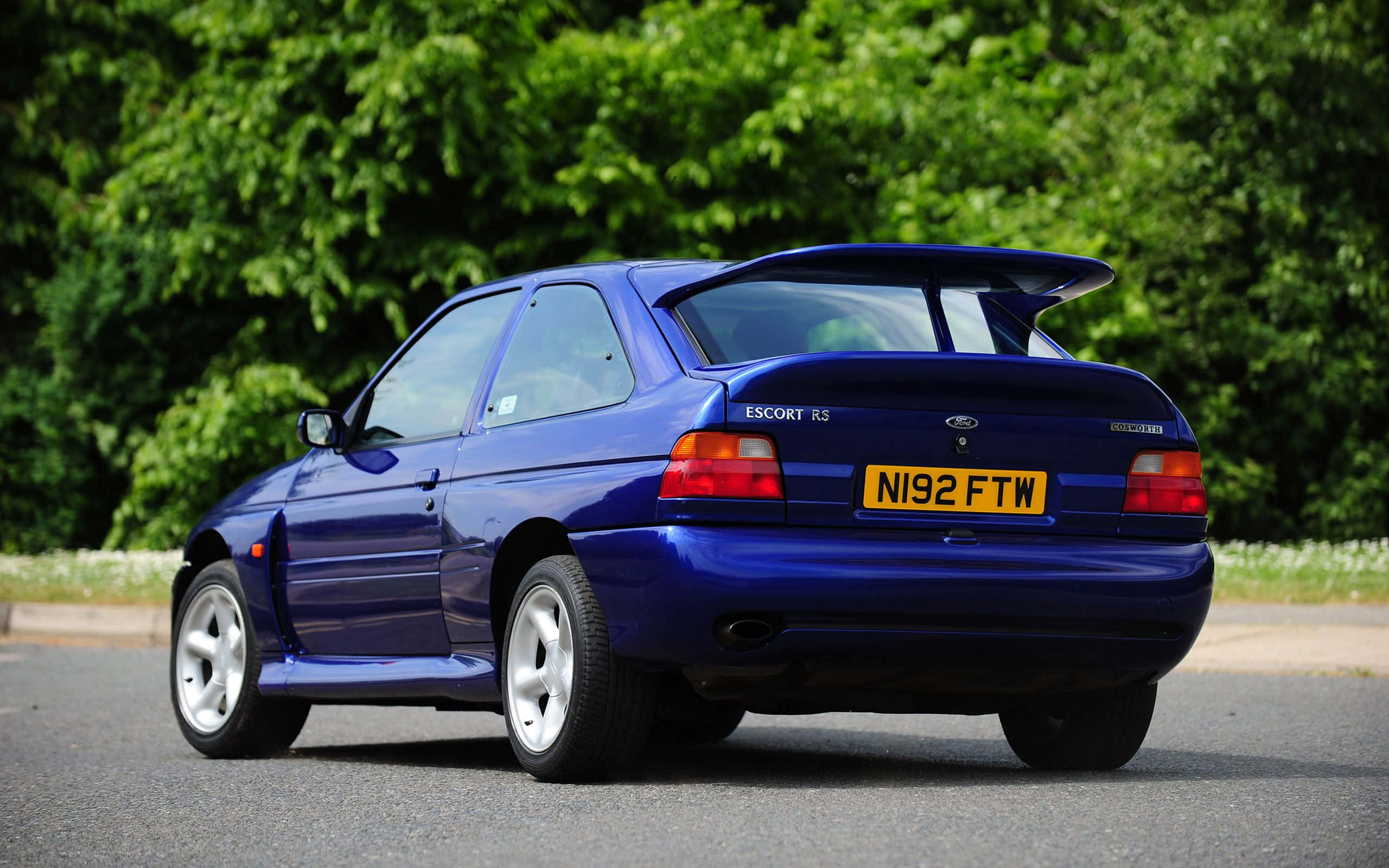 Ford Escort 桌布 壁紙 — 一輛充滿活力的藍色福特 Escort RS1800 跑車停在瀝青路上，背景是茂密的綠樹。