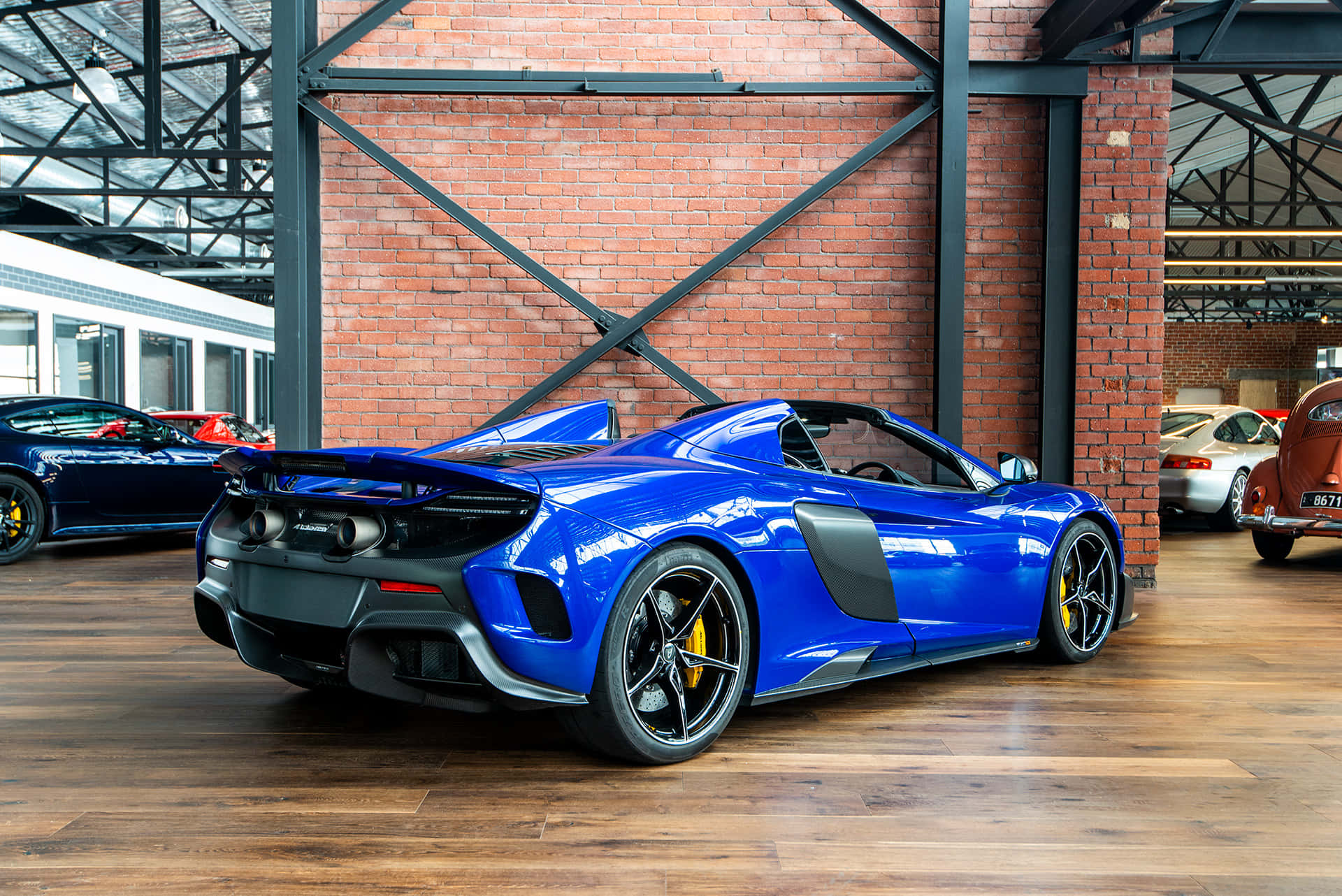 Mclaren 675lt Spider 桌布 壁紙 — 一輛充滿活力的藍色McLaren跑車在明亮的燈光下，在一個質樸的磚木車庫中與其他豪華車輛一起閃閃發光。