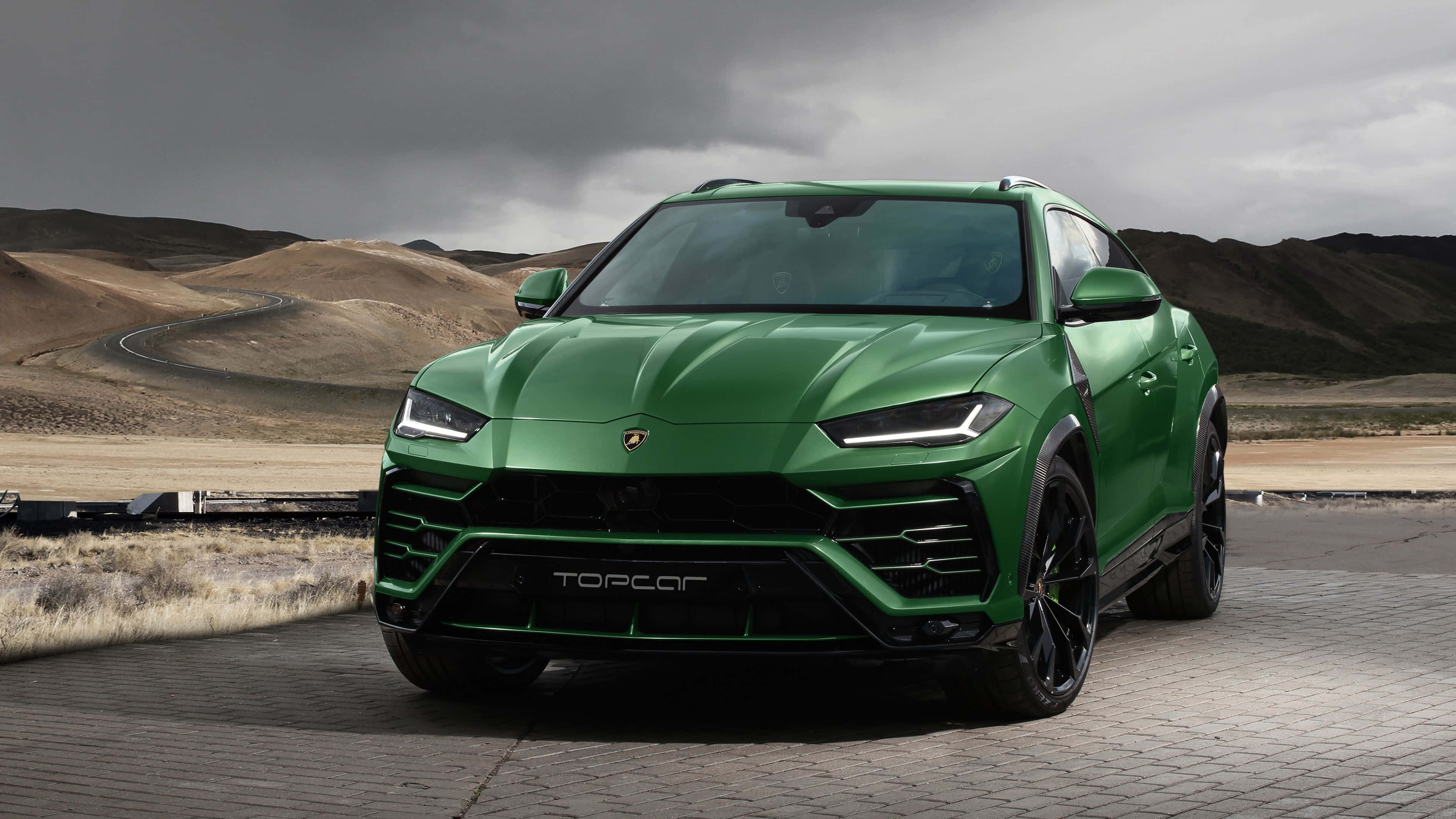Lamborghini Urus 桌布 壁紙 — 一輛充滿活力的綠色林寶堅尼 Urus Performante 停在鋪砌的觀景點上，背景是戲劇性的多雲天空和荒蕪的山丘。
