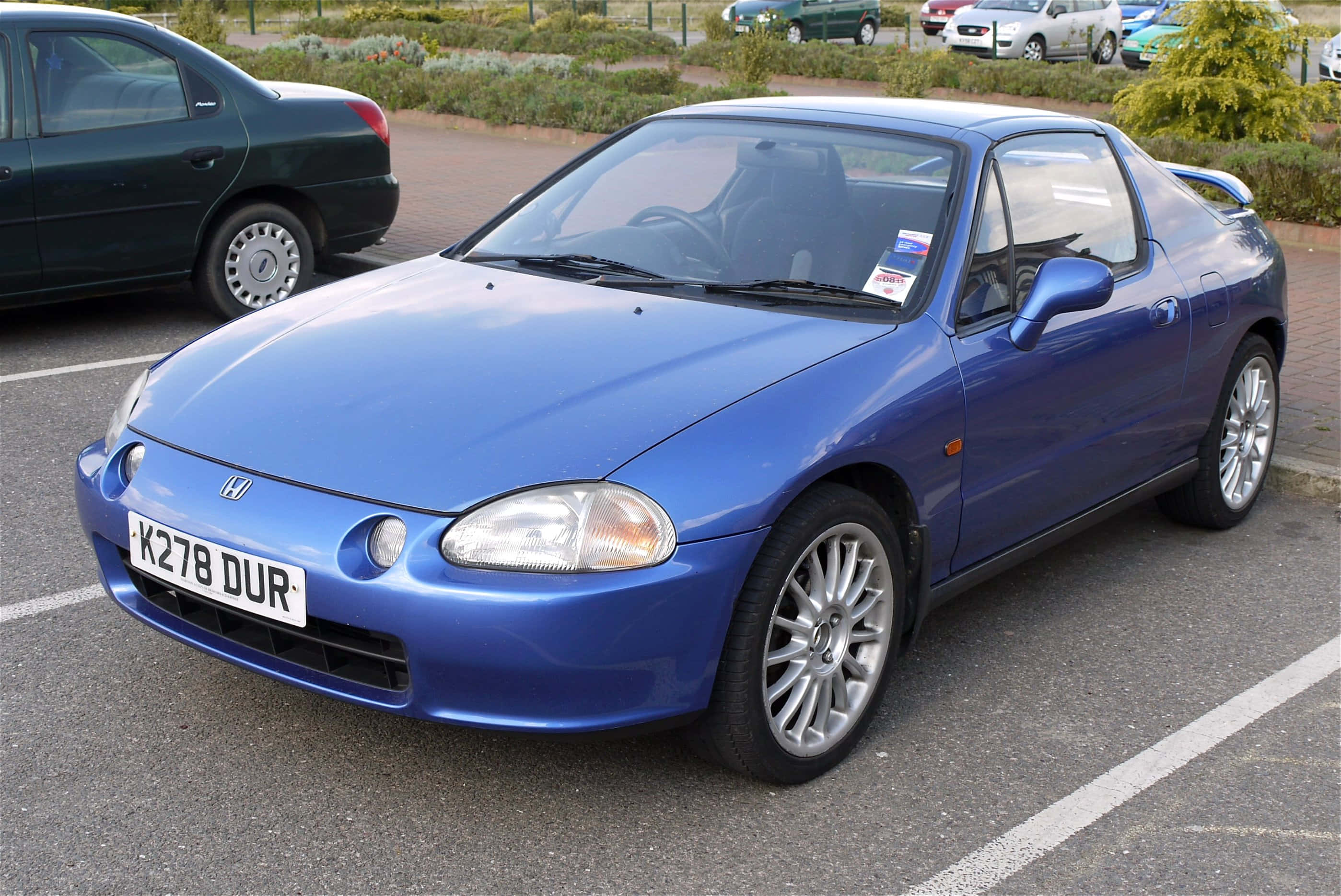 Honda Civic Del Sol 桌布 壁紙 — 一輛充滿活力的藍色本田 Del Sol 開篷車停放在日間停車場，背景有其他車輛和綠化。