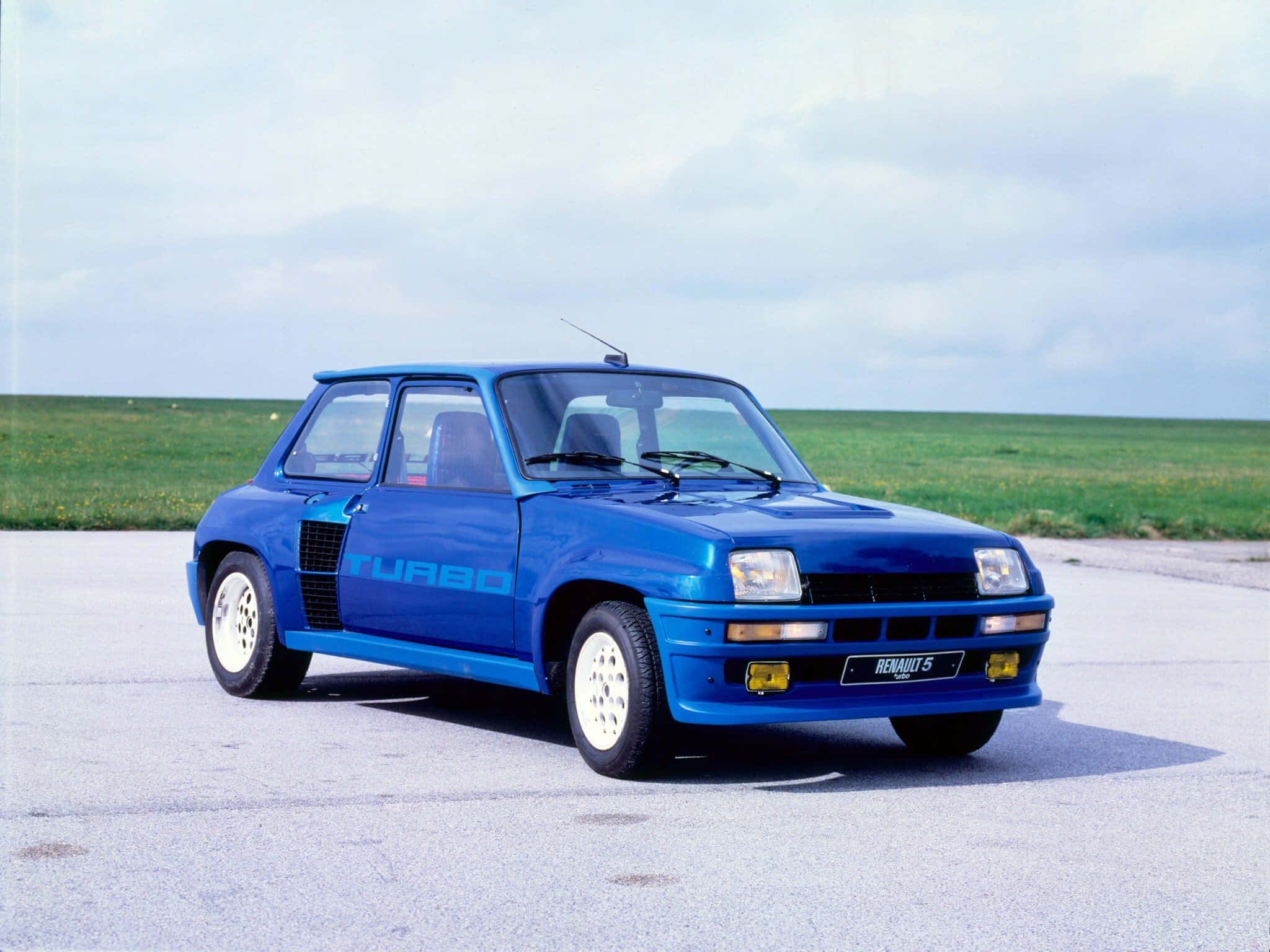 Renault 5 Turbo 桌布 壁紙 — 一輛充滿活力的藍色雷諾5 Turbo跑車停泊在鋪砌路面上，背後有草地，天空多雲。